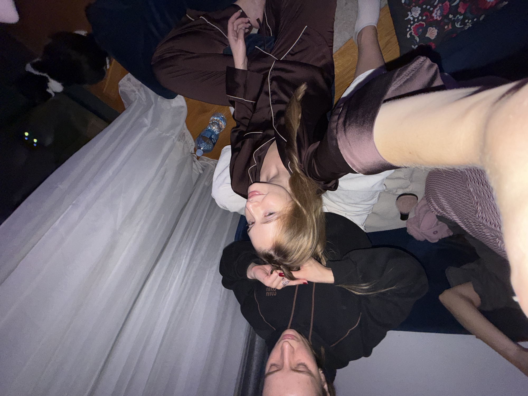 Two young women are lying on a bed or floor, presumably indoors. One woman wears a dark-colored sweatshirt with a logo on the front. The other is wearing brown pajamas with white piping. To their left, a water bottle sits next to a white curtain. A black and white cat is visible in the upper left corner of the frame. The image is a selfie. No identifiable details indicate the city of Belgrade, Serbia.