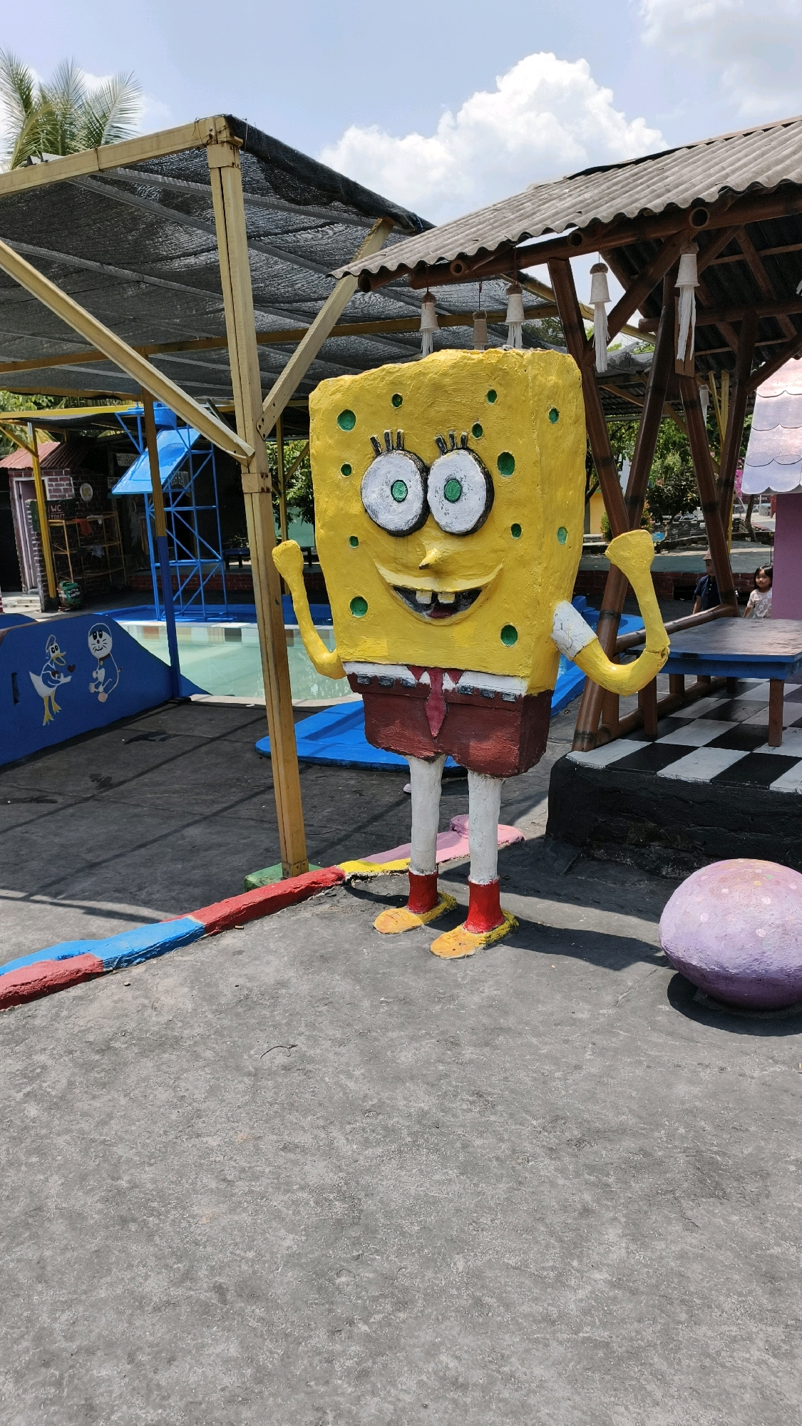 A life-sized, yellow Spongebob Squarepants statue stands on a grey, asphalt-like surface in a recreational area. The statue is brightly painted, with exaggerated features, including large green-dotted eyes, a wide smile, and red shorts. It wears a red tie and white socks with yellow shoes. In the background, a shallow swimming pool with blue and white tiling is visible, surrounded by a yellow wooden structure with a black mesh covering. Adjacent to the pool is a blue slide with cartoon characters painted on it, including a duck and Doraemon. Further back, a thatched-roof gazebo with wooden pillars and hanging decorations is present. To the right of the Spongebob statue, a black and white checkered table and a large, purple, round object, possibly a decorative stone, are visible. The scene is outdoors, under a bright sky with scattered clouds, suggesting a sunny day. Palm trees are visible in the distance. The overall environment appears to be a water park or a family-friendly outdoor attraction. This location is identified as being in the city of Air Paoh, Indonesia. There are no discernible people interacting with the statue in the frame, although a child is partially visible in the background near the gazebo.