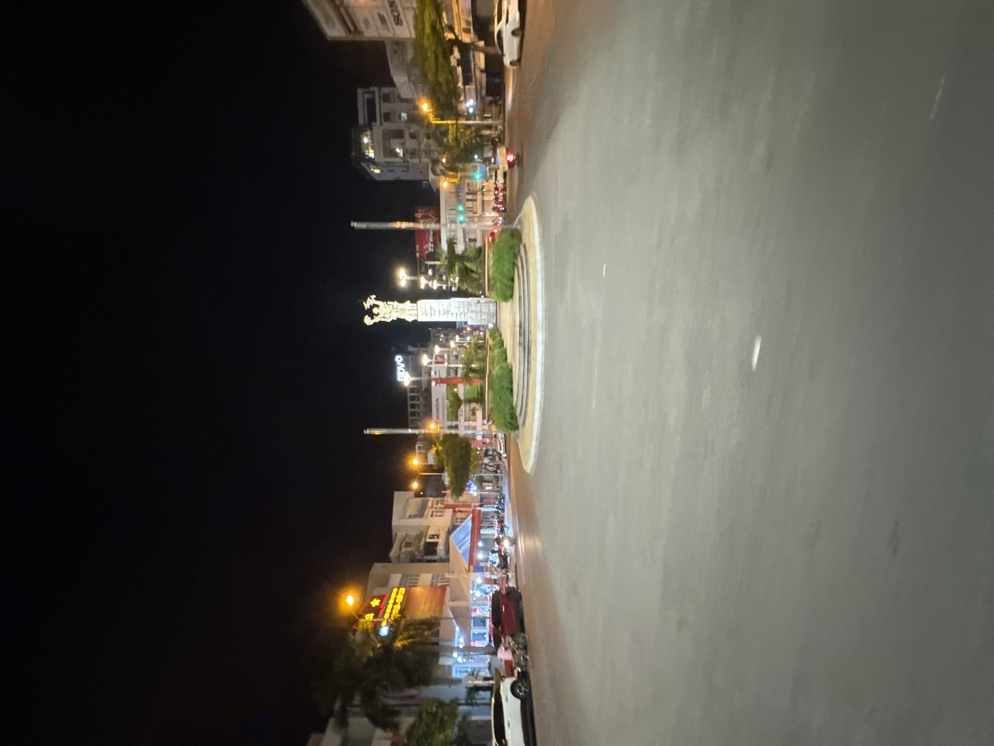 This image captures a nighttime view of a city roundabout in Nha Trang, Vietnam. The central focus is a monument featuring two figures atop a column. Surrounding the roundabout are various buildings, some illuminated with storefronts and signs, including what appears to be a "HONDA" dealership. Trees and greenery are visible along the edges of the roundabout and in front of the buildings. The street is paved and appears to be moderately busy with several cars and motorcycles in motion or parked. Streetlights and building lights illuminate the scene, casting a glow on the road and surrounding structures. The sky above is dark, indicative of nighttime.