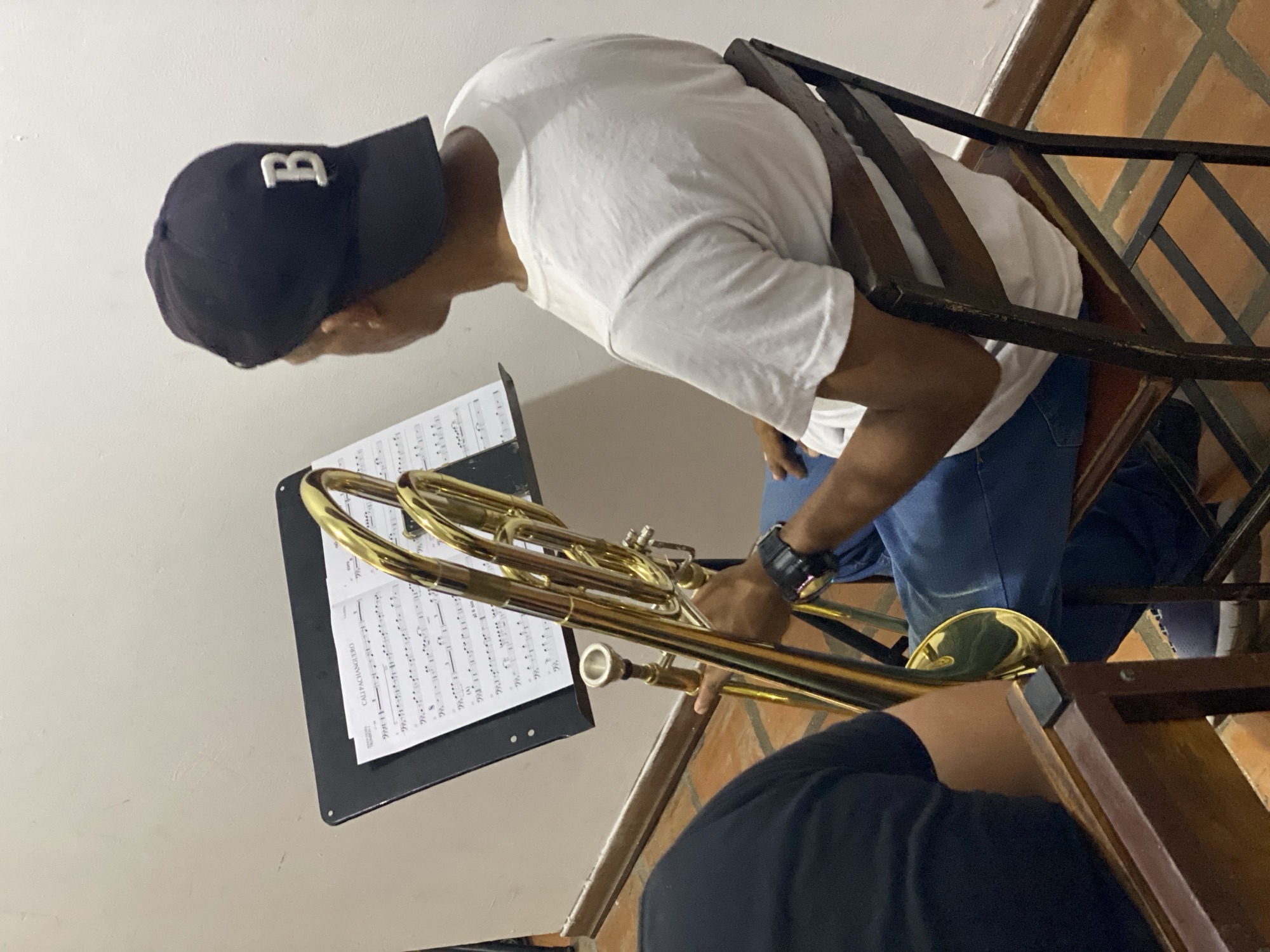 An indoor scene captures a musician playing a brass trombone. The primary subject is a person, appearing male, with dark skin, wearing a white short-sleeved t-shirt, blue jeans, and a dark baseball cap with a prominent white capital "B" on the front. A dark wristwatch is visible on his left wrist. He is seated on a dark wooden chair, seen partially, and leans forward, his head bowed, focused on the sheet music.
In front of the musician, a shiny gold-colored trombone is held in playing position, its various tubes and slides gleaming. A black music stand holds sheet music, displaying musical notation and the title "CALI PACHANGUERO" clearly visible at the top left of the page.
The setting appears to be an interior space, possibly a home or a practice area in Tinaquillo, Venezuela. The background features a light-colored wall and a distinctive floor made of reddish-brown and dark square tiles arranged in a geometric pattern, possibly indicating a staircase or an elevated landing. A dark wooden railing or balustrade runs diagonally across the image. A partial view of another person's arm, dressed in black, is visible in the lower foreground. The lighting suggests an indoor environment. The musician exhibits clear focus and concentration on his instrument and the score.