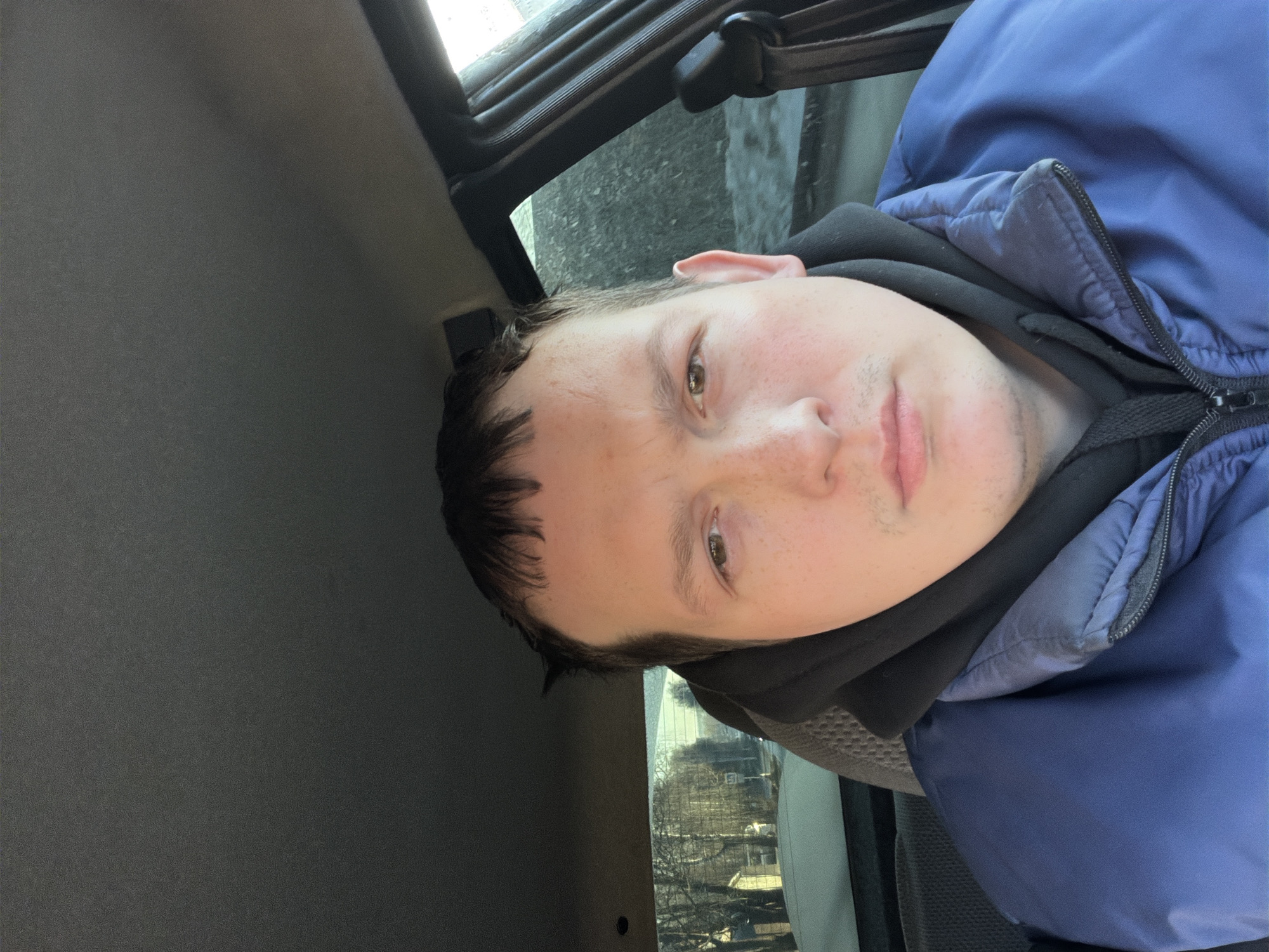 A close-up selfie of a person with short brown hair and fair skin, wearing a black hooded sweatshirt underneath a blue padded jacket. The person appears to be inside a vehicle with a dark grey interior. The vehicle's window shows a blurred outdoor scene, revealing bare trees and buildings, which are suggestive of a cityscape. This could potentially be the city of Chaikovsky, Russia. The scene is taken from a low angle, looking up at the person.
