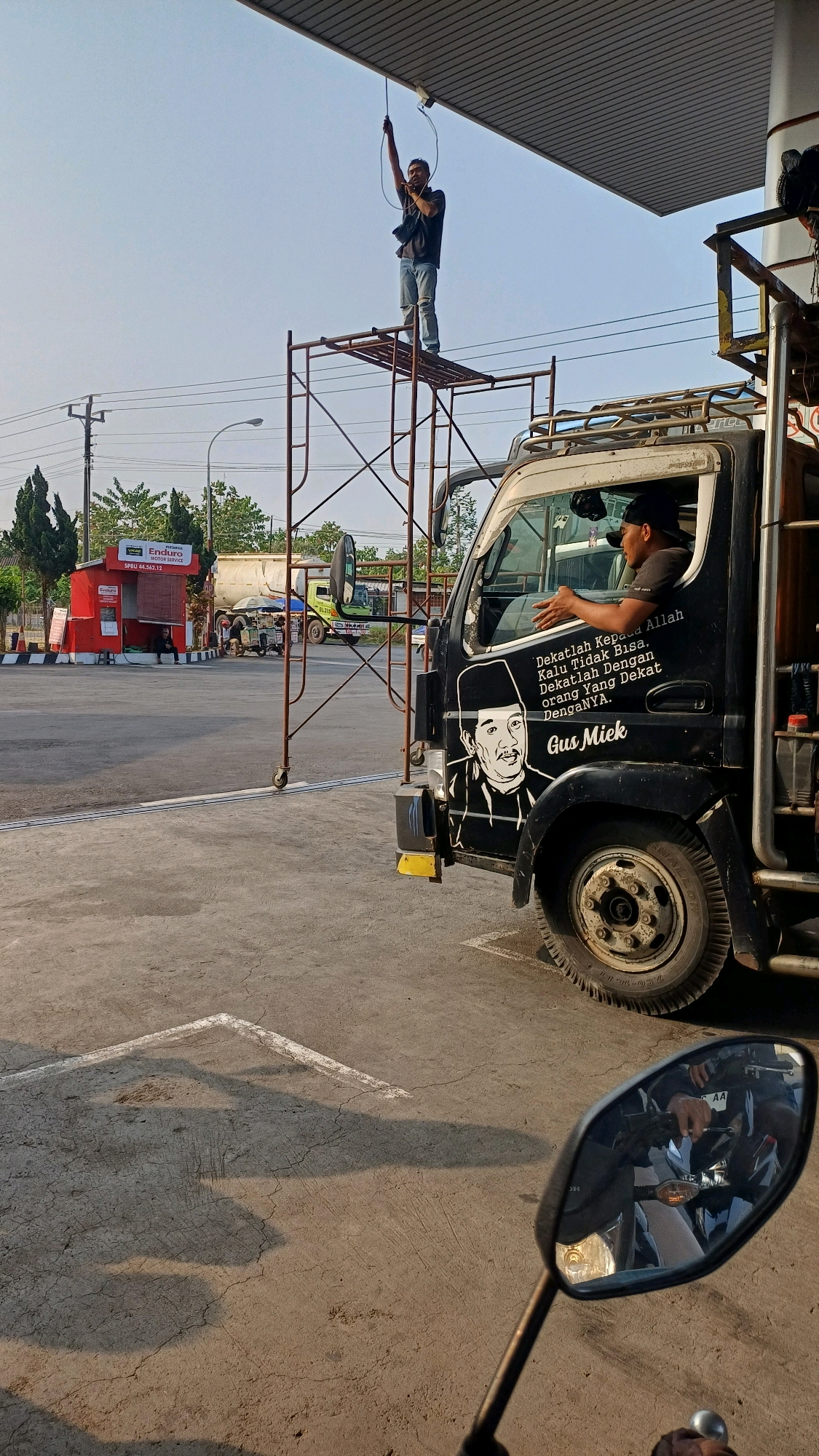 A man stands on scaffolding, holding a wire, under the roof of a gas station in Kranggan, Indonesia. A truck with a portrait of Gus Mick and text that reads "Dekatlah Kepada Allah Kalau Tidak Bisa Dekatlah Dengan Orang Yang Dekat Dengannya." is parked nearby. The driver of the truck is visible through the window, looking out. A motorcycle is reflected in the truck's side mirror, showing the rider's hands on the handlebars. The scene is set on a concrete surface with white line markings, suggesting a parking or service area. Trees and other vehicles are visible in the background, indicating a public area. The lighting suggests it is either early morning or late afternoon.