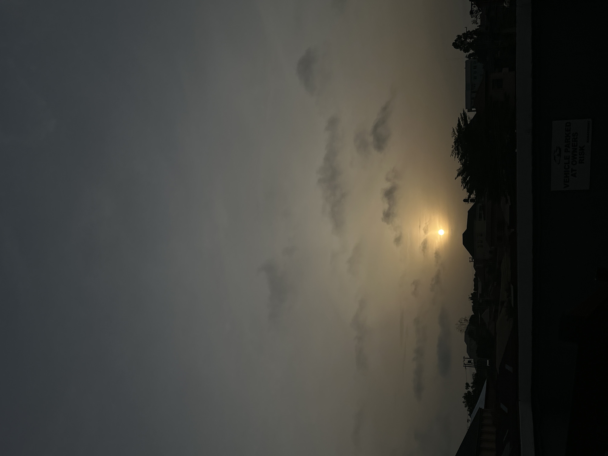 The image captures a sunset scene in Jos, Nigeria. The sky is filled with clouds, casting a grey-yellow hue across the landscape. The sun is visible as a bright orb amidst the clouds. In the foreground, there is a building and trees silhouetted against the sky. A sign is seen near the building with the inscription "VEHICLE PARKED AT OWNERS RISK." The scene is generally dusky, with the details of the buildings and landscape partially obscured by the low light.
