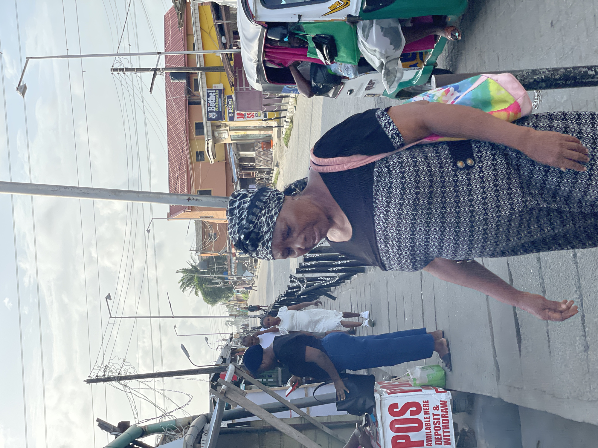The image captures a bustling street scene in Ikorodu, Nigeria, during the daytime under an overcast sky.
In the foreground, an older woman with dark skin is prominently featured, looking slightly downwards. She wears a black and white patterned dress, a matching headscarf, and has a pink strap visible over her shoulder, attaching to a colorful, tie-dye pattern bag. Her right arm is extended forward, with her hand open as if in a gesture of asking or offering.
Behind her, on a paved sidewalk, several other individuals are visible. One person in a black top and blue pants is standing near a white box-like structure displaying a "POS AVAILABLE HERE DEPOSIT & WITHDRAW" sign, suggesting a mobile banking or payment point. Further down the sidewalk, another person is dressed in white.
The background reveals a typical urban street with commercial buildings. One prominent building has a yellow facade and a reddish roof, featuring a sign that reads "Bethel Schools." Above the street, numerous utility poles are visible, entangled with a dense network of power lines and electrical wires. Streetlights are also mounted on some of these poles. A white three-wheeled vehicle, commonly known as a Keke Napep or auto-rickshaw in Nigeria, is partially visible parked on the side of the road, displaying a "NIKE" logo. Distant palm trees hint at the tropical climate. The overall scene depicts daily life and commercial activity in the area.