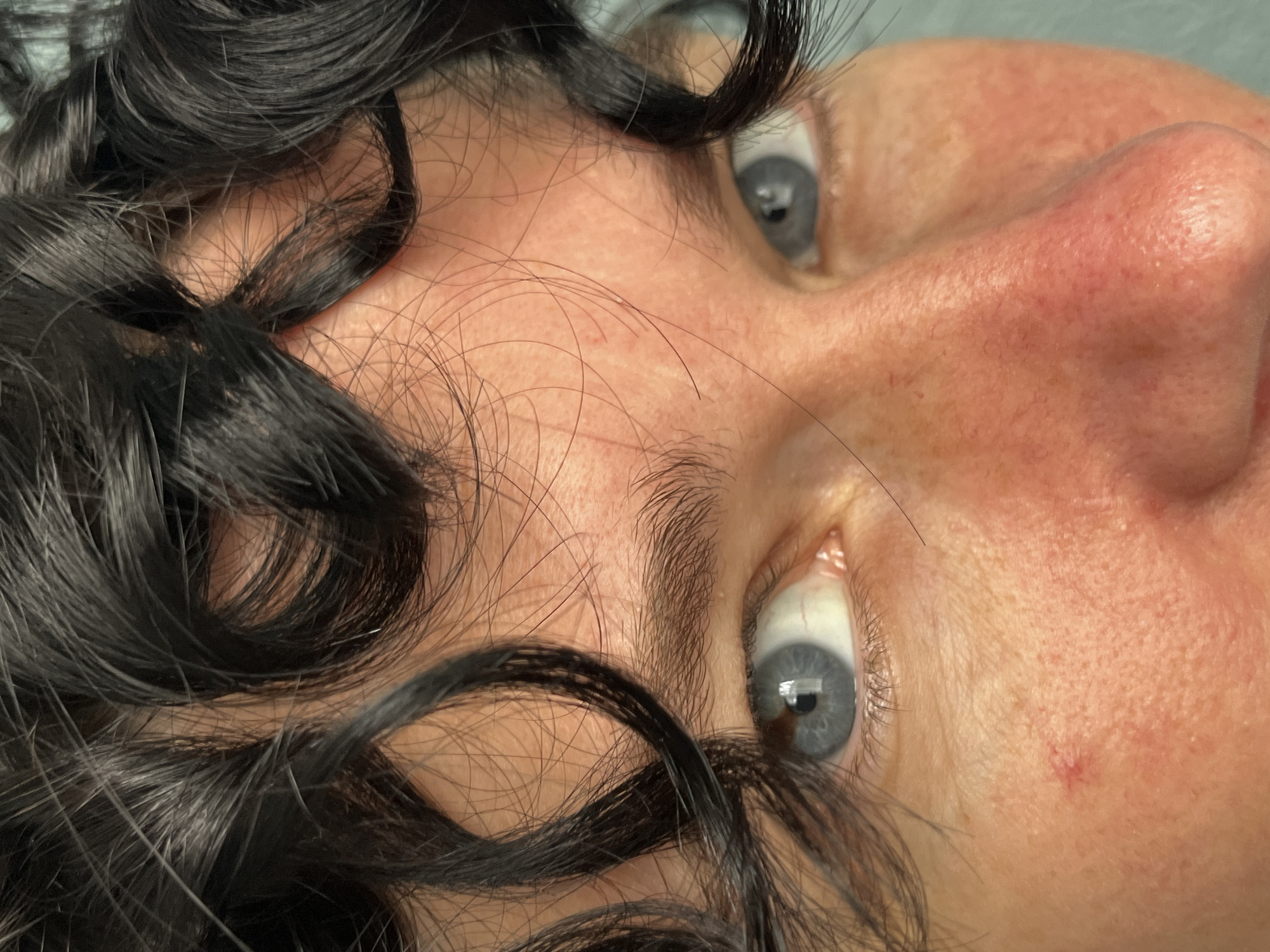 The image is a close-up, eye-level shot of a person's face, focusing on their eyes, nose, and forehead. The subject has dark, curly hair framing their face, with strands falling over their forehead and near their eyes. Their eyes are a striking light blue, and their eyebrows are dark and well-defined. The skin on their face is fair with some redness and visible pores, particularly around the nose. A small red mark is visible on the left cheek. The background appears to be a solid, light blue surface, suggesting an indoor setting, possibly a studio or a plain wall. There is no discernible location in the city of ღრმაღელის თემი, Georgia, as the image is a close-up of a person's face with no contextual background elements.