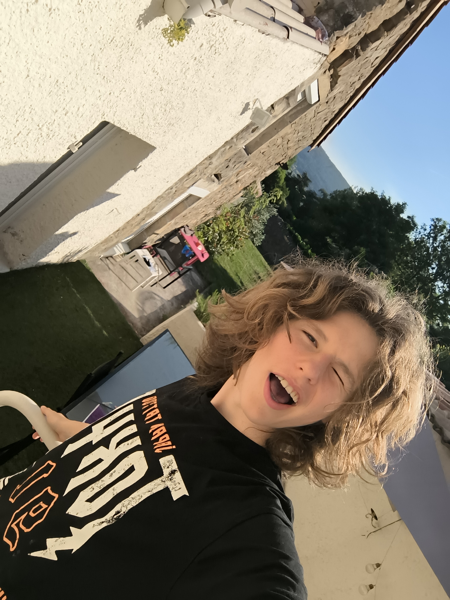 A young person with wavy, light-brown hair wearing a black t-shirt with white and orange graphics is captured in a selfie outdoors. The individual is smiling with an open mouth and one eye closed in a wink. The background shows a mix of architectural and natural elements. A textured, white stucco wall with a window is visible, as well as a stone structure along the roofline. In the distance, there's greenery, trees, and what appears to be a distant mountain range under a clear blue sky. Based on the location, it is likely the setting is a residential area in Pélussin, France. There is a lawn, an outdoor table and chairs, and a child’s toy set, indicating the presence of a home and family. A white chair or railing is in the lower left corner.