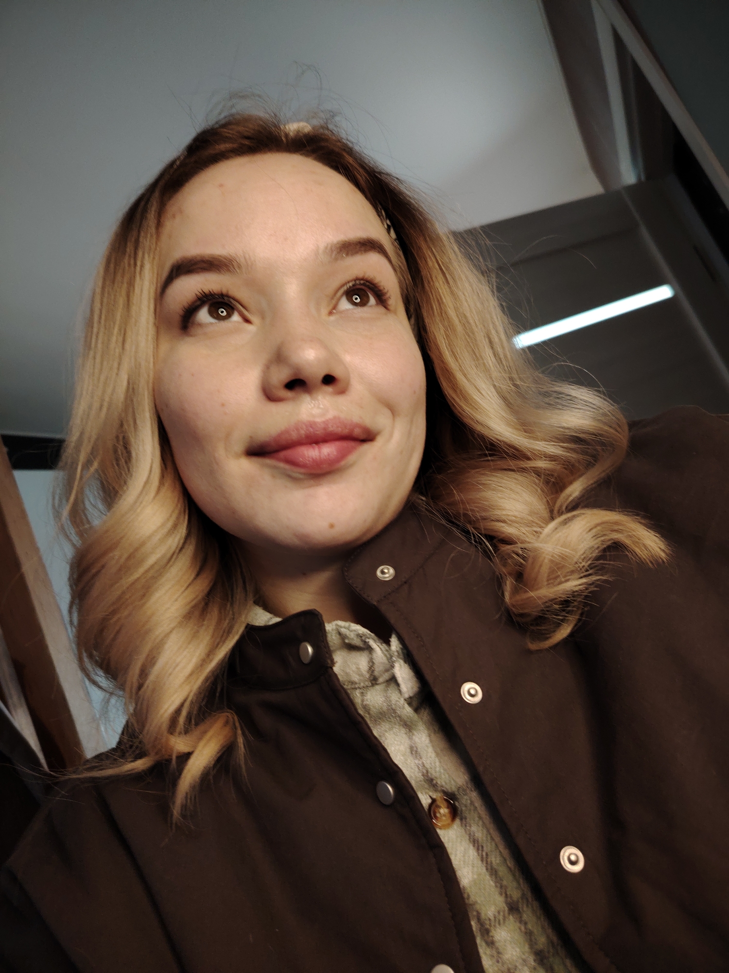 A low-angle close-up captures a young woman with blonde, wavy hair. She is looking up and to the right. Her skin has a few visible freckles, and she has full lips. She is wearing a brown jacket with silver snap closures over a plaid shirt. The background appears to be an indoor setting, with a white ceiling and a linear light fixture visible. The location in the city of Марковский, Russia, cannot be determined from this image.