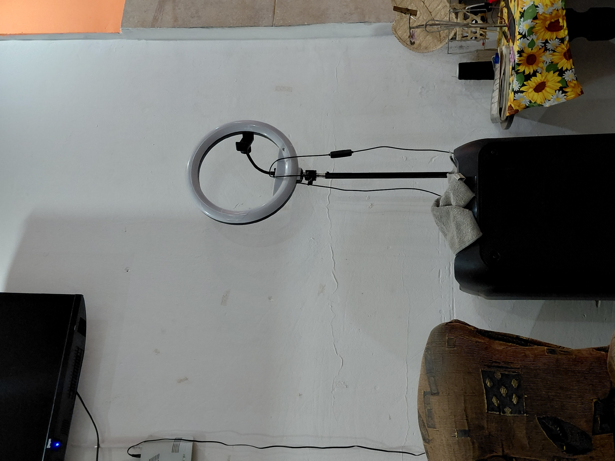 The image, oriented horizontally, captures a section of an indoor room with a prominent white wall showing signs of wear and texture.
In the center, a white circular ring light is mounted on a black stand, with multiple black cables connecting to it and extending towards the right. These cables lead to a black electronic device, likely a speaker or soundbar, positioned on the right side of the frame, with a light-colored towel draped over its top. Above this device, a colorful fabric patterned with sunflowers and daisies hangs, partially obscuring a light-colored, possibly woven or wooden, decorative object.
Towards the bottom right, part of a distinctively patterned, possibly woven or wooden, piece of furniture is visible. In the bottom left quadrant, a black monitor or television screen is angled, displaying a small blue light indicating it is powered on. Faint text, possibly "Sveon," is discernible on the monitor's lower frame. Below the monitor, a white modem or router is situated, from which several black cables extend and run along the lower portion of the wall.
The upper left corner of the room features a patch of wall painted in an orange color, contrasting with the dominant white. The ceiling in the upper right appears to be made of light-colored tiles or wood. The scene suggests a functional, possibly cluttered, interior space, likely used for multimedia activities or content creation, given the presence of the ring light. No people are visible, and no specific activity is occurring. The location context is Santa Ana, Venezuela.