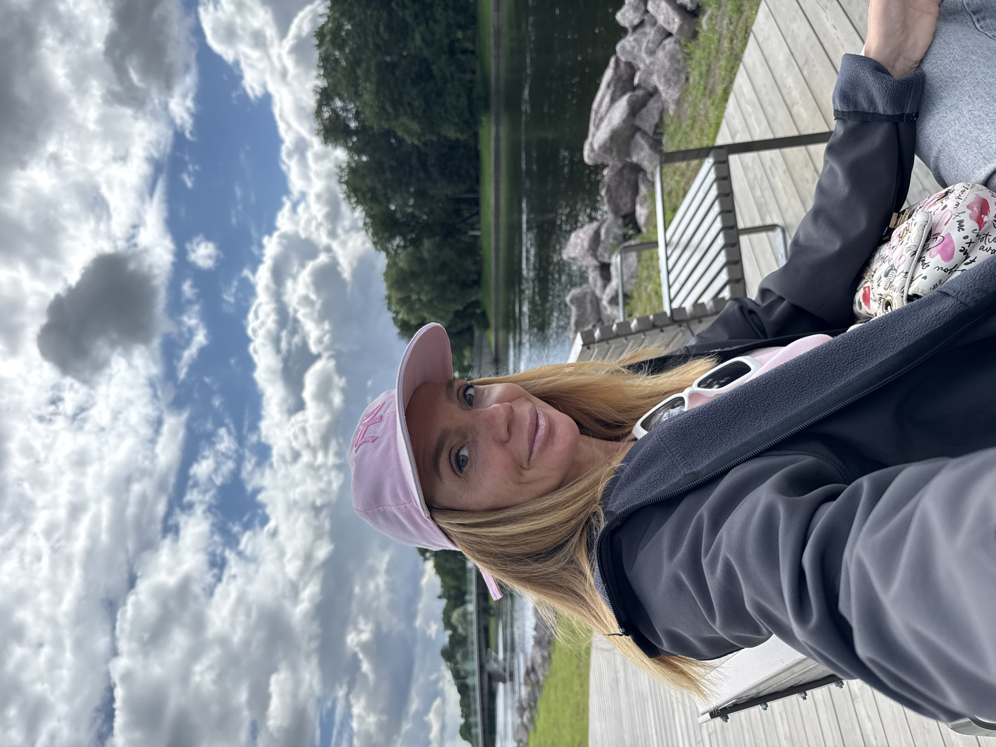 A woman wearing a pink baseball cap and a dark jacket is seated on a wooden deck next to a body of water. She is looking at the camera and smiling. Behind her, there is a river with a bridge in the distance and trees lining the opposite bank. The sky is cloudy. This scene is located on the waterfront in Saint Petersburg, Russia.