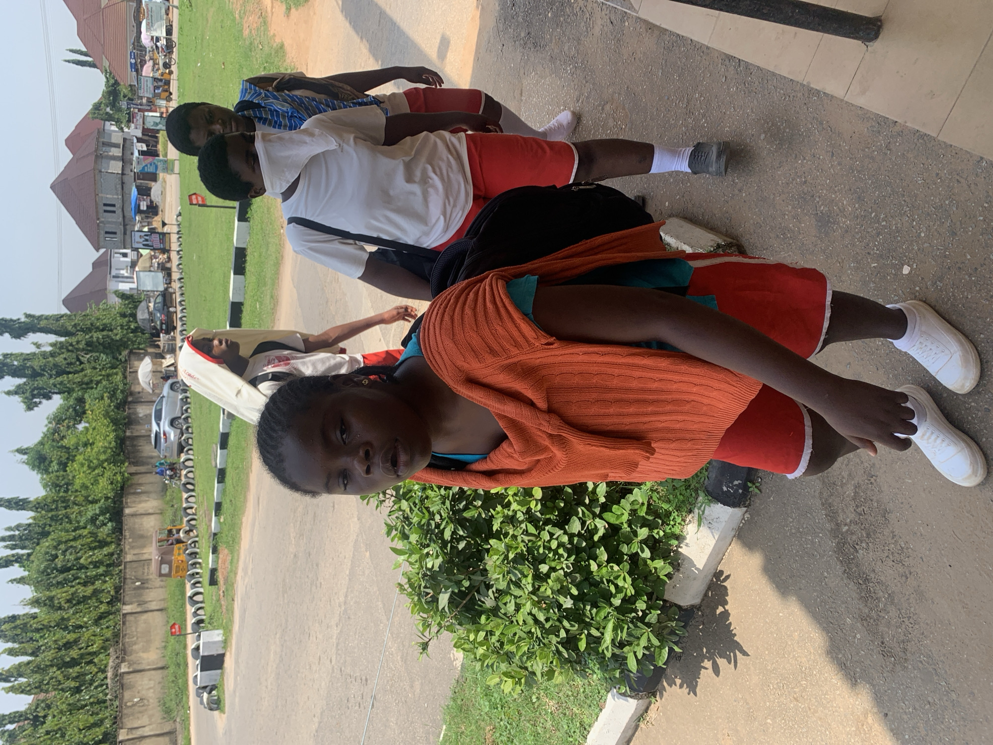 In Abuja, Nigeria, a group of four young girls walks along a paved path next to a grassy median bordered by tires. The lead girl, wearing an orange knitted vest over a blue shirt and red shorts, is looking towards the camera. To her right, a second girl is partially obscured, wearing a white t-shirt, red shorts, and a white cloth covering her mouth and nose. Behind her, a third girl, in a blue and white striped t-shirt and red shorts, is also partially visible. The fourth girl, in the background, is wearing a white hooded garment and red shorts. The environment includes a street with a car and a tricycle, buildings with red roofs in the distance, and a concrete wall with trees behind it. The lighting suggests a sunny day.