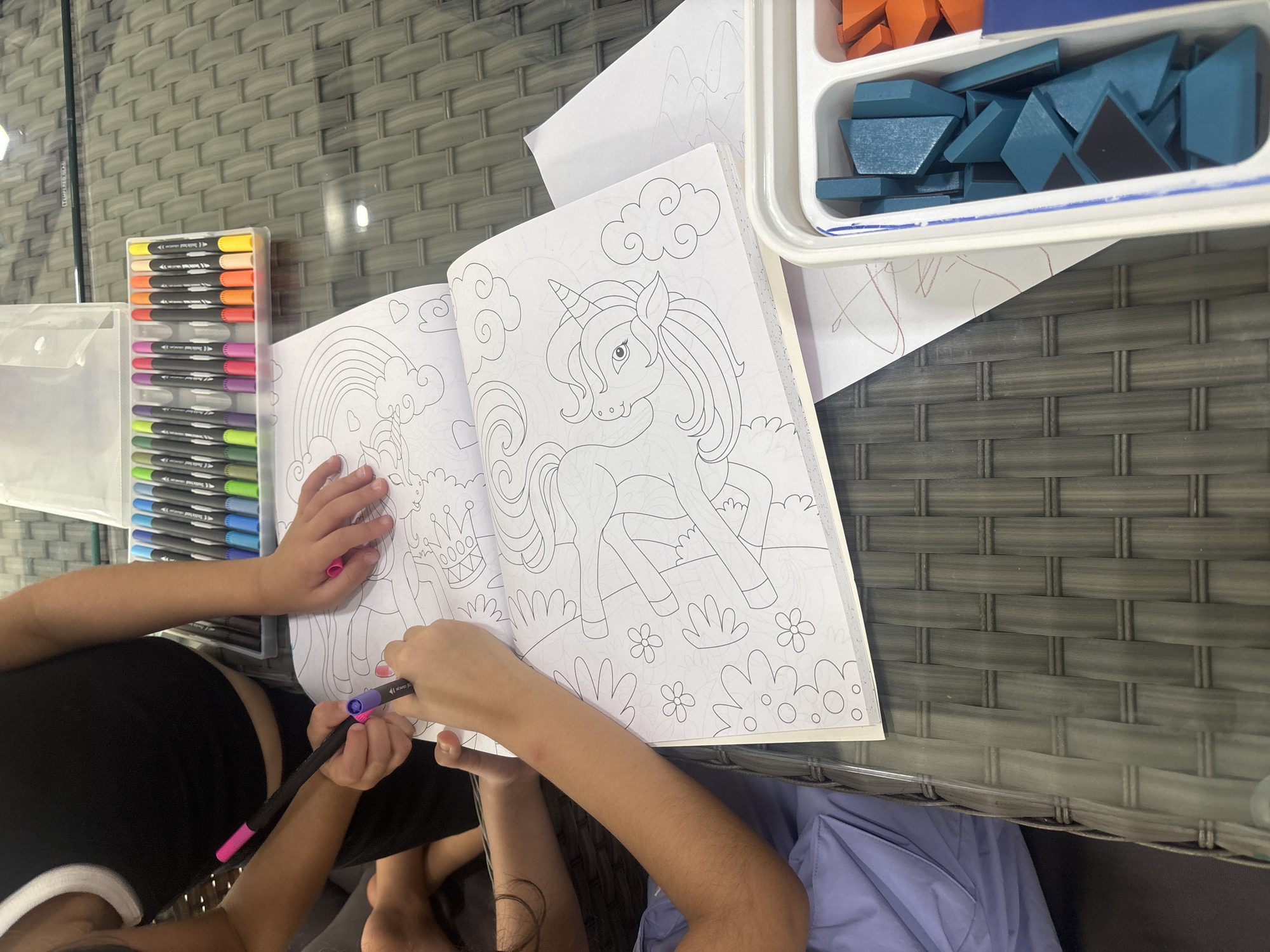 In Dubai, United Arab Emirates, two individuals are engaged in a creative activity at an outdoor table. The table, made of woven grey material and topped with glass, displays an open coloring book featuring unicorn and rainbow illustrations. A set of multi-colored markers is neatly arranged in a clear plastic case to the right of the book. One individual, wearing a black tank top with a white trim, is using a purple marker to color the book. Their hands are positioned over the coloring book, and a pink marker cap is visible. The other individual's arms are also visible, suggesting they are also participating in the activity. To the right of the coloring book, a white plastic container holds an assortment of teal and orange geometric wooden blocks, possibly for a separate activity or game. The scene is lit by natural daylight.