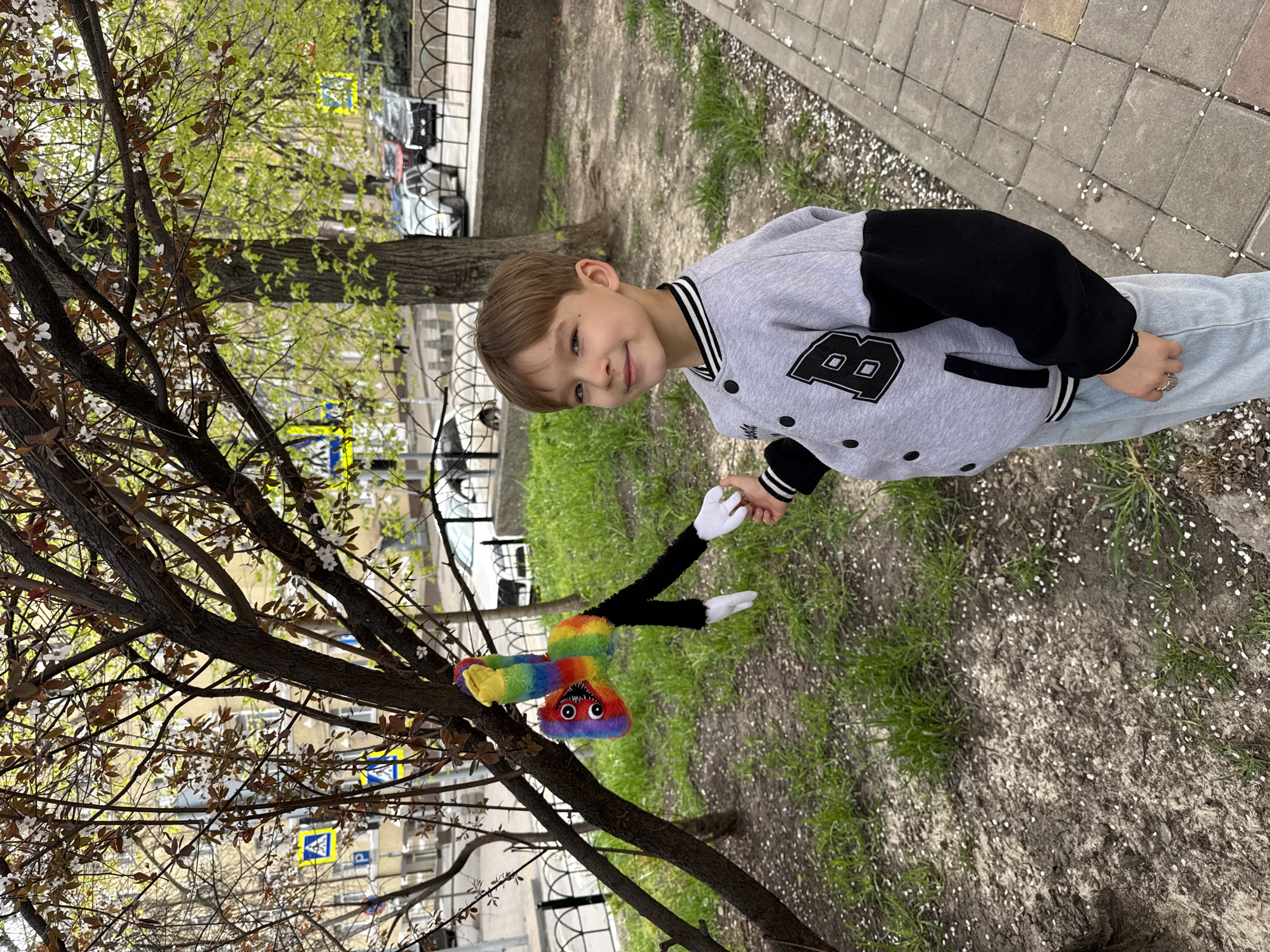 A young boy stands near a tree on a city street, possibly in Rostov-on-Don, Russia. He is wearing a grey varsity jacket with black sleeves and the letter "B," light blue jeans, and has short brown hair. He smiles and looks towards a colorful plush toy that appears to be stuck on a branch of the tree. The toy has a rainbow-colored body, large eyes, a red mouth with sharp teeth, and black legs with white feet. In the background, there is a green patch of grass, a sidewalk paved with grey tiles, a metal fence, and buildings, along with parked cars. A yellow road sign with a blue pedestrian crossing symbol is visible. The tree has bare branches interspersed with small white blossoms, and there are fallen petals on the ground.