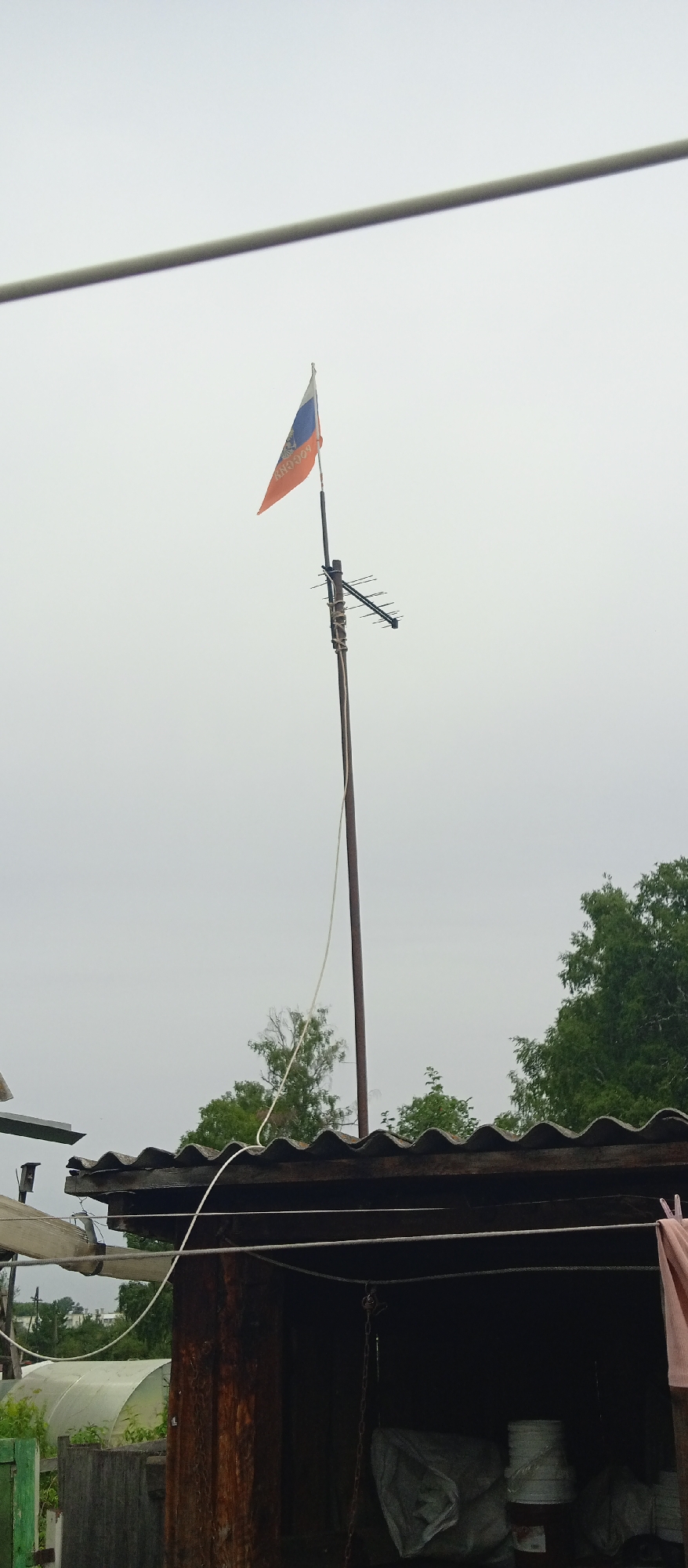 A Russian flag, orange with blue and white stripes, is affixed to a metal pole. The pole also supports a television antenna. A white rope is tied to the pole and extends downwards, passing over a corrugated roof of a wooden structure. The structure appears to be an open-sided shed or garage, with some items visible inside, including buckets and covered objects. Trees are visible in the background. The sky is overcast. The scene is consistent with a residential area in Nazarovo, Russia.