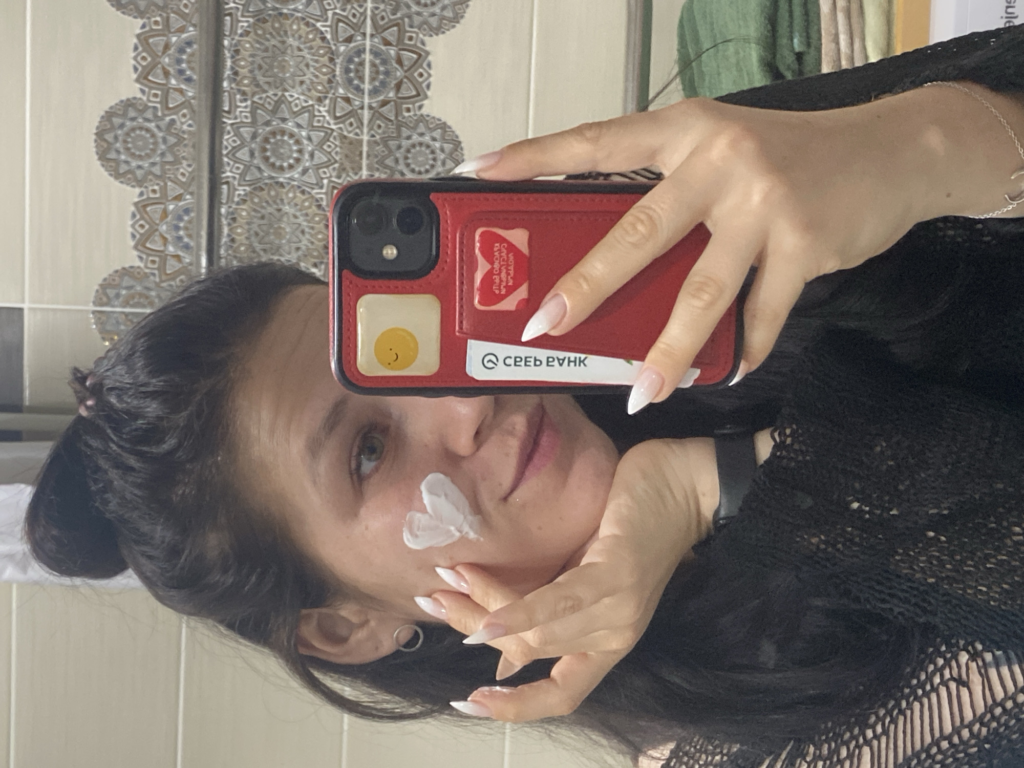 A woman is taking a selfie in a bathroom with patterned tiles. She has dark hair pulled back in a bun and a daub of white cream on her cheek. She's holding a red iPhone in her left hand, which has a picture of a fried egg on the back and a card with the words "СБЕР БАНК" visible. She has long, white-tipped fingernails. She wears a black, mesh-like top. The environment consists of the bathroom's patterned tile background. The location is Chaikovsky, Russia.