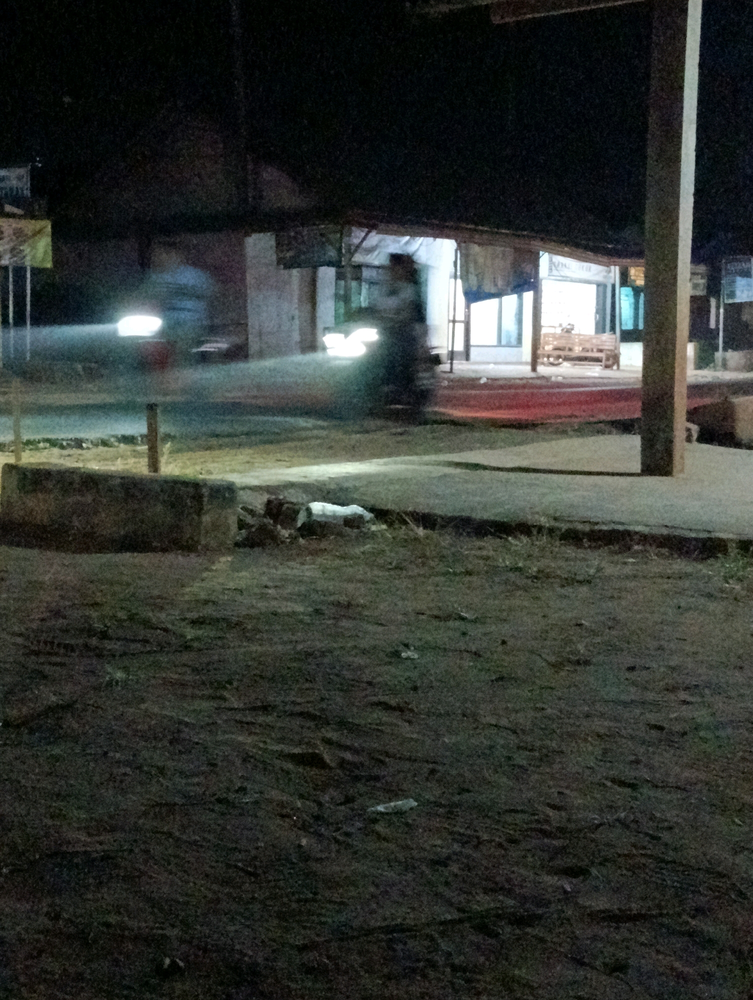 The image depicts an outdoor scene at night, likely in Adirejo, Indonesia. In the foreground, there is a patch of dark, uneven ground with some scattered debris. A concrete structure, possibly a low wall or planter, is visible on the left. The middle ground features a paved area with a person on a motorcycle, indicated by the blurred motion and bright headlights. Behind the motorcycle, a building with a covered awning is visible, featuring windows and a wooden bench outside. The building appears to be a shop or a small establishment. The overall lighting is dim, with the headlights of the motorcycle and the lights from the building being the primary sources of illumination. A thick vertical post, likely part of a larger structure, stands on the right side of the image. The background is dark, suggesting open space or more distant structures. The scene appears to be a roadside or a public area, possibly a commercial strip, at night.