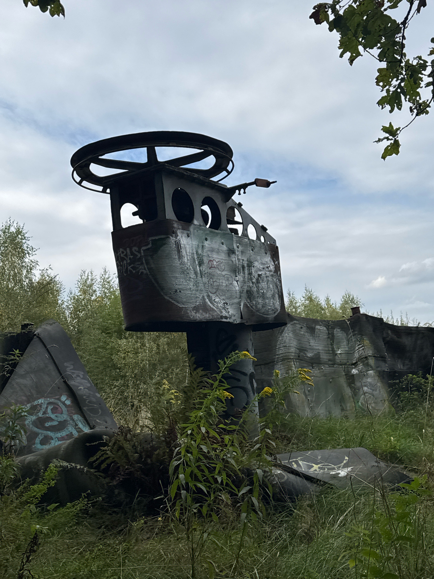 The image captures an outdoor scene featuring elements of a derelict industrial or military structure, possibly part of a bunker or defense system, situated amidst overgrown vegetation. The primary subject is a tall, cylindrical metal structure with a large circular wheel or mechanism at its apex. This structure is covered in graffiti. To the right, a long, sloped concrete wall, also bearing graffiti, extends into the background, partially obscured by trees. In the foreground, various pieces of metallic debris, likely also part of the structure, lie scattered among tall grass, ferns, and yellow wildflowers. The environment appears wild and untamed, suggesting neglect. The sky above is overcast with scattered clouds. The location is identified as Katowice, Poland. There are no people visible in the image.