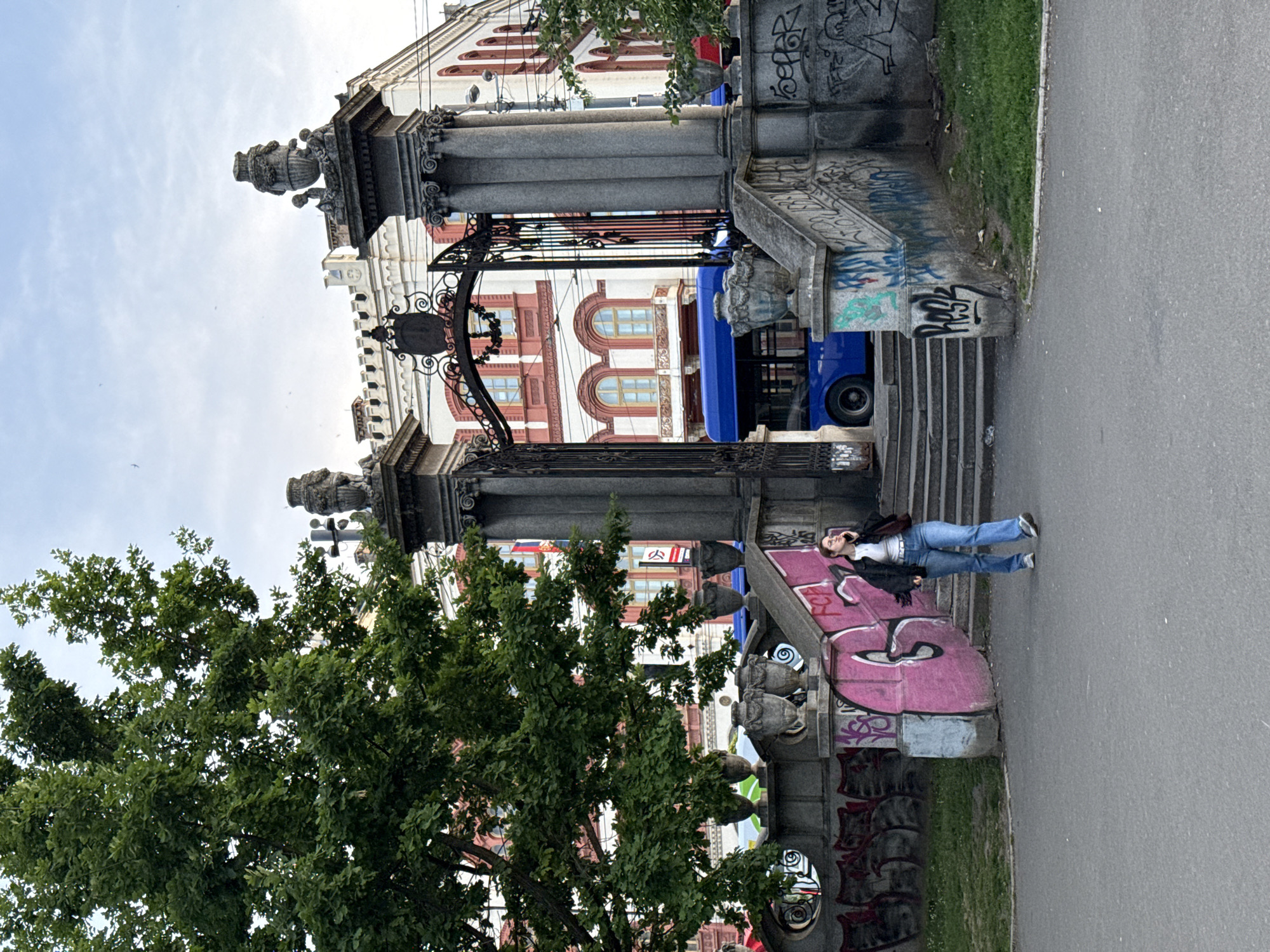 A street scene features an ornate stone gate with columns, decorative urns, and ironwork. Graffiti covers parts of the structure. A woman in jeans and a jacket is lying on a stone ledge beside the gate, seemingly on her phone. A tree with dense foliage is on the left side of the frame. In the background, a building with a red and white facade is visible behind the gate. A blue bus is parked behind the structure. The scene appears to be in Belgrade, Serbia.