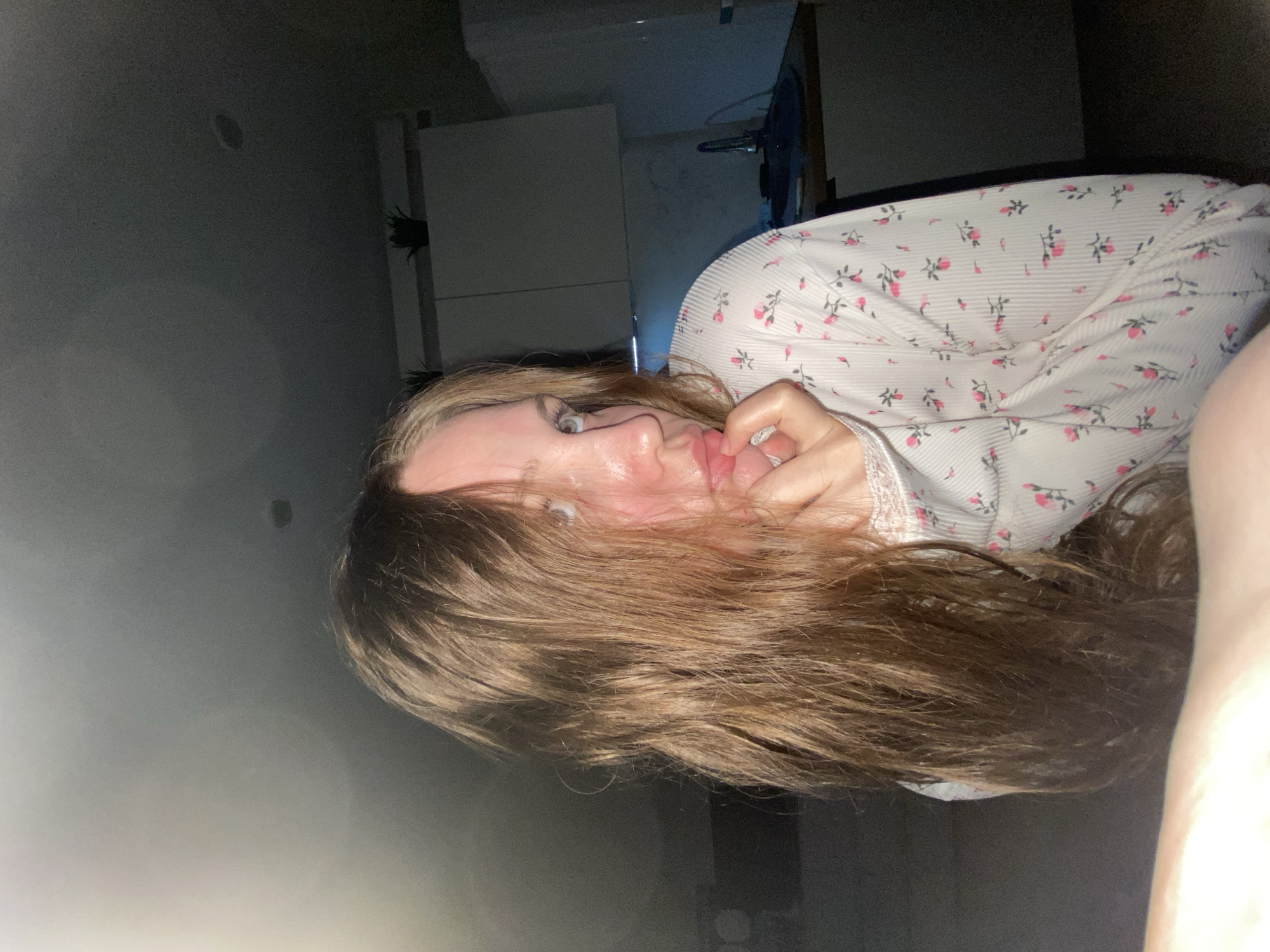 A woman is lying down, possibly in a bedroom, with her head close to the camera. She has brown hair and is wearing a white nightgown with a pink floral pattern. Her hand is near her mouth. The background includes a wall and a glimpse of furniture. There is no information to place the location in the city of Izhevsk, Russia.