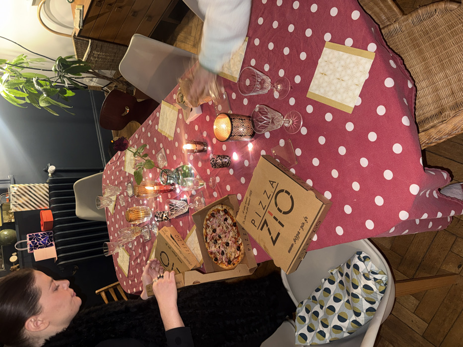 A tilted aerial view shows a table set with a red and white polka dot tablecloth, glasses, candles, and place settings. Two opened pizza boxes from "Pizza Zio" sit on the table, one with a pizza inside. A woman in a black outfit is seated at the table, and another person is partially visible at the top of the frame. A plant and decorative items are in the background.