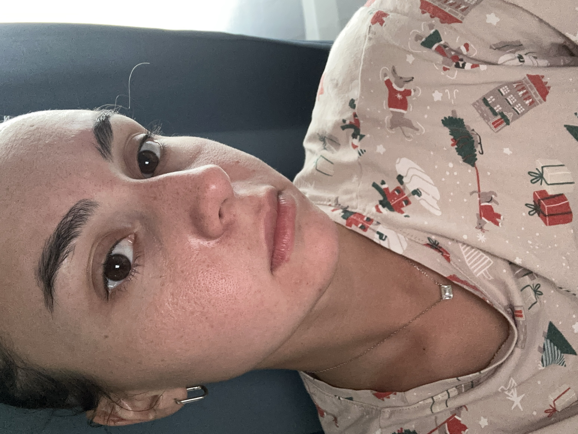 A close-up, rotated portrait shows a person, likely female, looking directly at the camera with a neutral expression. The image is taken indoors, possibly in Trudovaya, Russia. The individual has dark eyes and thick, dark eyebrows. Their skin displays natural texture with visible pores and some freckling, and appears slightly moisturized. A few light, wispy hairs are visible above the forehead.
The person is wearing a light beige top or pajamas featuring a festive Christmas pattern. The pattern includes cartoon depictions of reindeer in red sweaters, Santa Claus, Christmas trees, gift boxes, and white snowflakes. A delicate, silver-colored chain necklace with a small, clear, rectangular pendant is visible at the base of the neck. A distinctive silver-colored earring, shaped like an open safety pin, is worn in the left earlobe.
The background consists of a soft, dark blue or gray surface, suggesting a comfortable, relaxed setting like a sofa or bed. The lighting is soft and even, highlighting the subject's features. No visible text is present in the image. The attire indicates a casual, perhaps holiday-themed, indoor atmosphere.