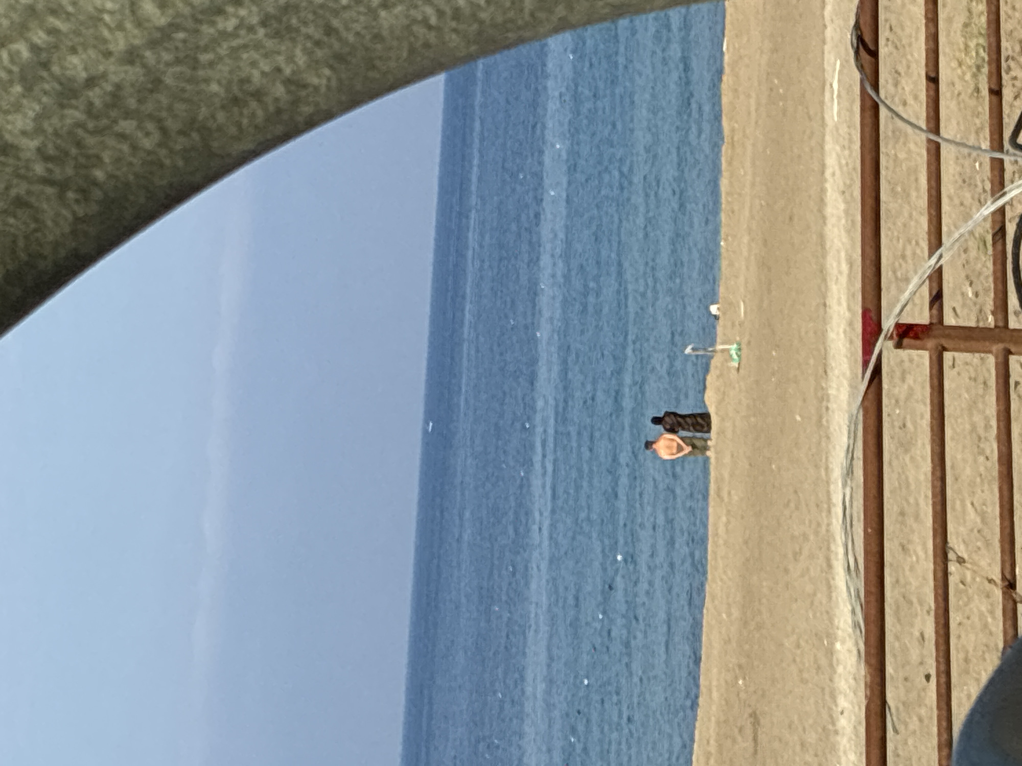 Two figures stand on a sandy beach facing the blue ocean. One figure, dressed in a brown garment, stands to the right of another figure, who is wearing an orange shirt and green shorts. The ocean stretches to the horizon, meeting a pale blue sky. A large, curved, grey object obscures the upper right portion of the image. In the foreground, a rusty metal railing runs horizontally across the bottom of the frame. A thin strand of wire is draped over the railing. The beach has scattered debris and a white object that resembles a marker or buoy is visible in the sand. The scene is devoid of any identifiable landmarks or structures that would place it specifically in Alanya, Turkey.