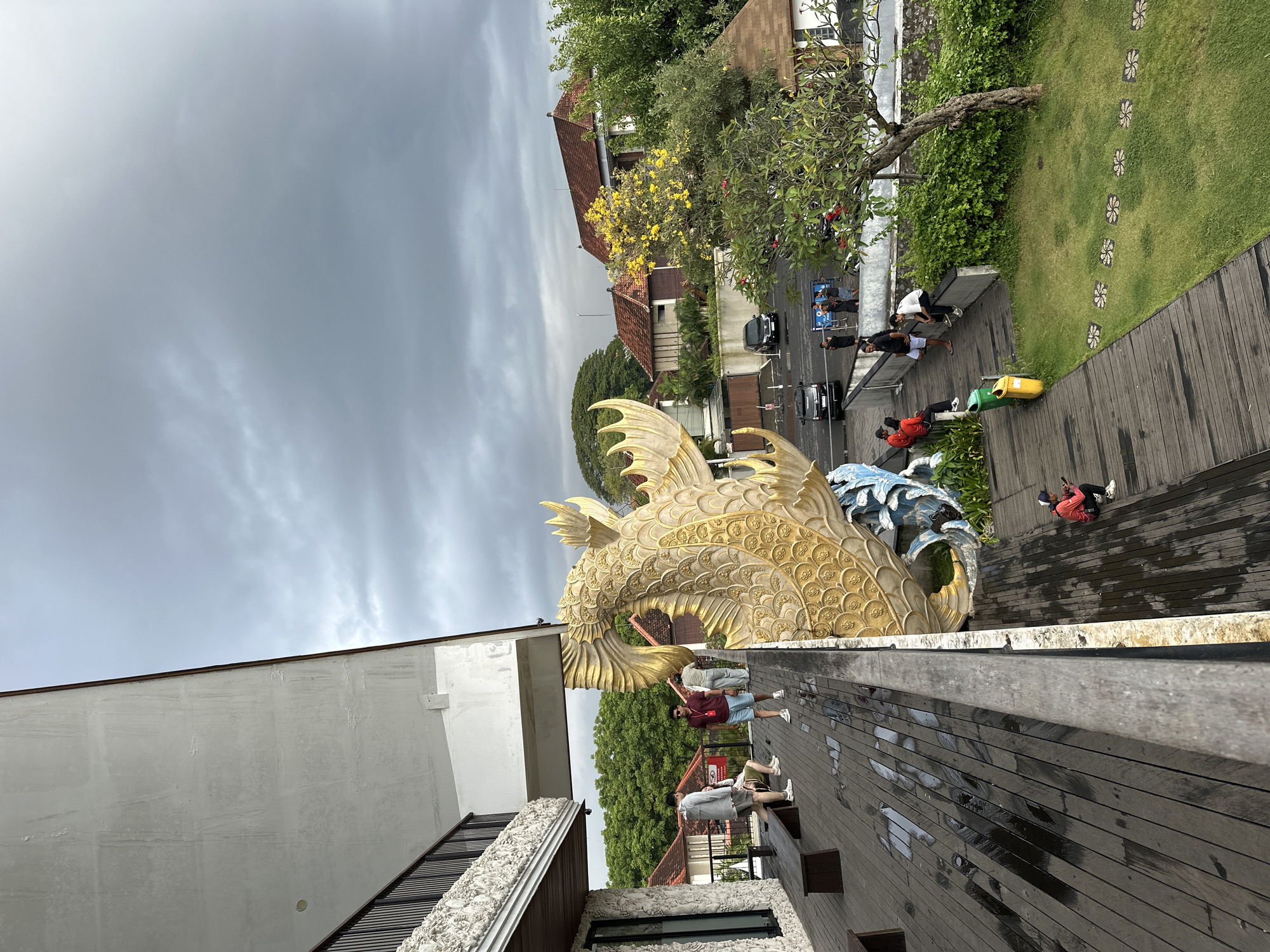 The image shows a scene in Denpasar, Indonesia, featuring a large, golden dragon statue alongside a wooden deck area. The dragon is intricately detailed with scales and fins. On the deck, several people are standing, walking, and sitting, some of whom appear to be tourists. The background includes a street scene with cars, other people, and traditional Balinese architecture. Vegetation is present, including trees and manicured grass. The sky is overcast. The perspective suggests the photo was taken from a higher vantage point, potentially from a building.
