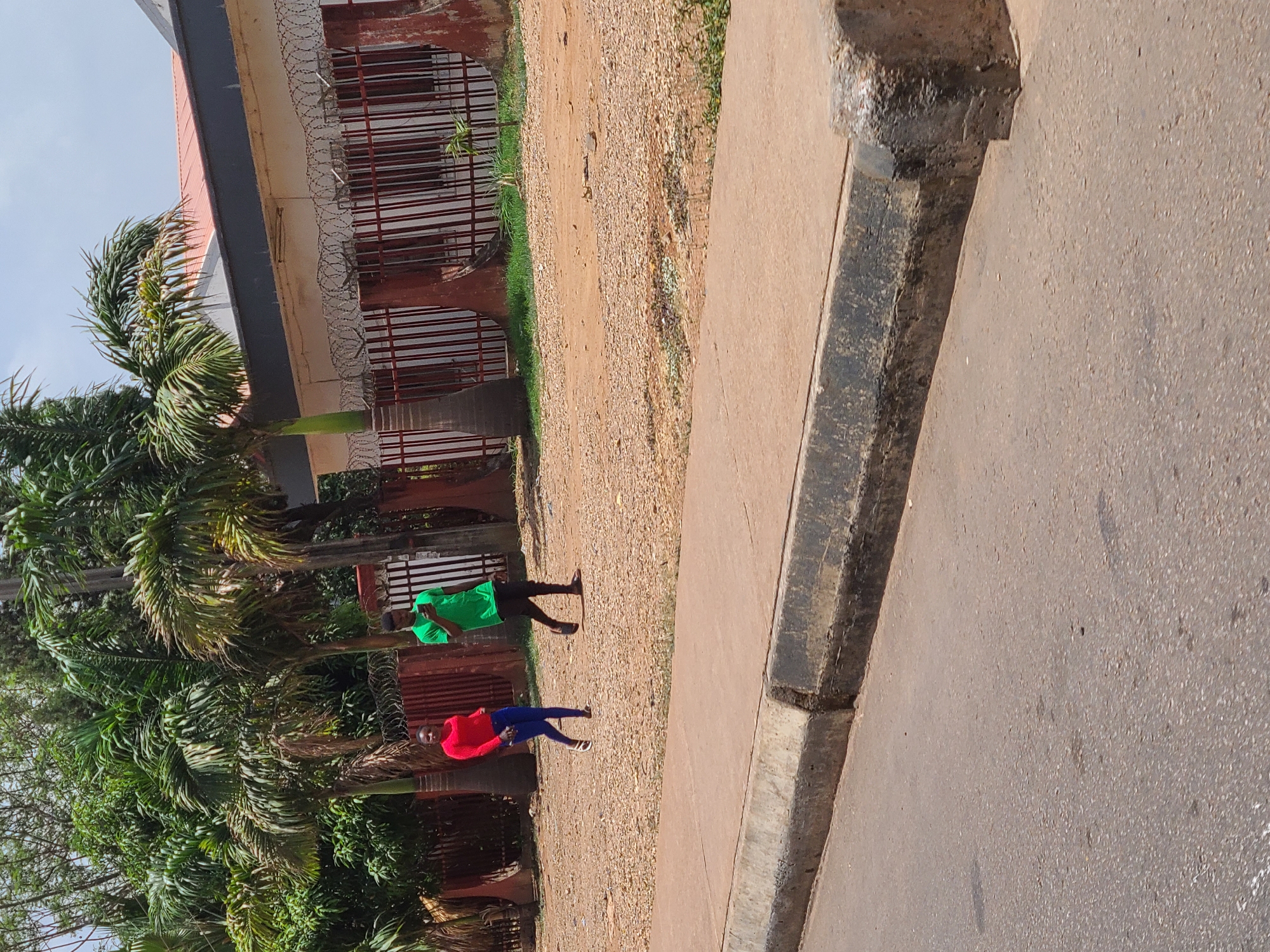 A daytime outdoor shot shows two people walking along a dirt path adjacent to a sidewalk and a building with a fence in Jos, Nigeria. The building has red window bars and barbed wire along the top of the fence. Lush green trees, including palm trees, are on the left side of the path. The person closer to the camera is wearing a red top and blue pants, while the person further away is wearing a green top and black pants. The sidewalk is made of concrete with a dark gray curb. The street is made of asphalt. The scene appears to be taken from a low angle and slightly rotated.
