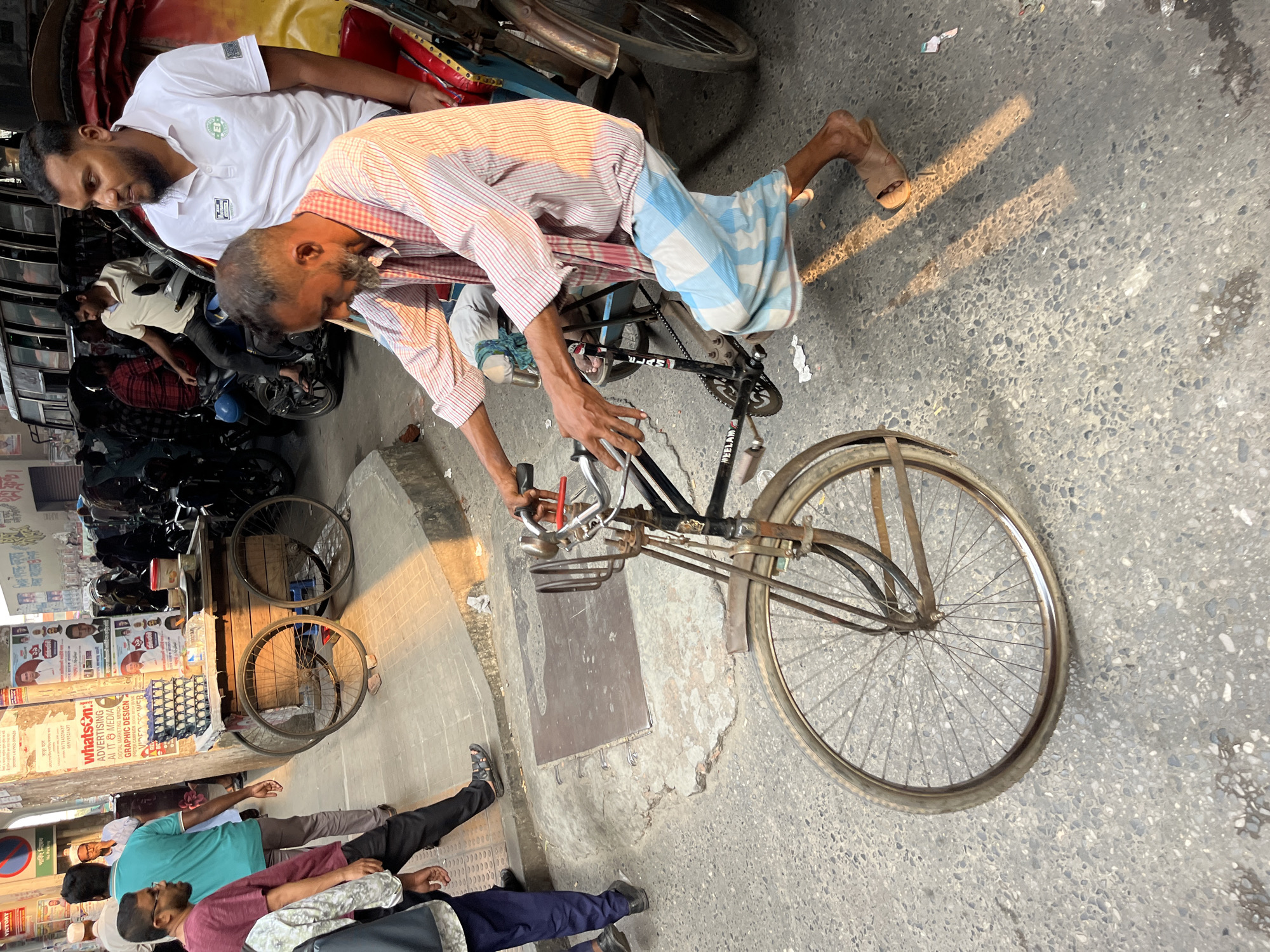 The image captures a street scene in Dhaka, Bangladesh. A middle-aged man wearing a striped pink and white shirt and a blue and white checkered sarong is bent over an old black bicycle, possibly fixing it or making an adjustment. The bicycle has a rusty front carrier and faded "MEELAM" branding on the frame. Another man in a white polo shirt with a small emblem is resting on the seat of a red rickshaw or auto-rickshaw nearby.
In the background, to the left, there is a row of motorcycles parked along the side of a building. A street vendor's stall or shop with advertising posters and stacked goods, including a stack of white rectangular objects and a blue carton, is also visible. Some men wearing t-shirts are seen on the left, lying on the ground beside the advertisements. The street surface is paved with concrete or asphalt, with evident wear and tear and patches. A metal access panel is integrated into the pavement near the man working on the bicycle. Signs reading "No Parking" and advertisements are also noted on the side of the building. The scene takes place in daylight, with light casting shadows on the street.
