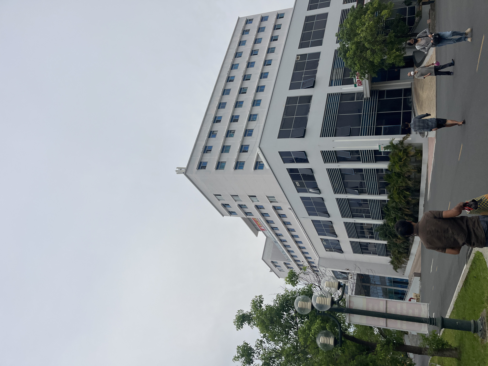 The image shows an exterior view of a tall, white building in Nha Trang Ward, Vietnam. The building has a modern design with many windows, some of which are open. There are people walking on the sidewalk beside the building. A row of trees and streetlights are in the foreground. The sky is overcast.