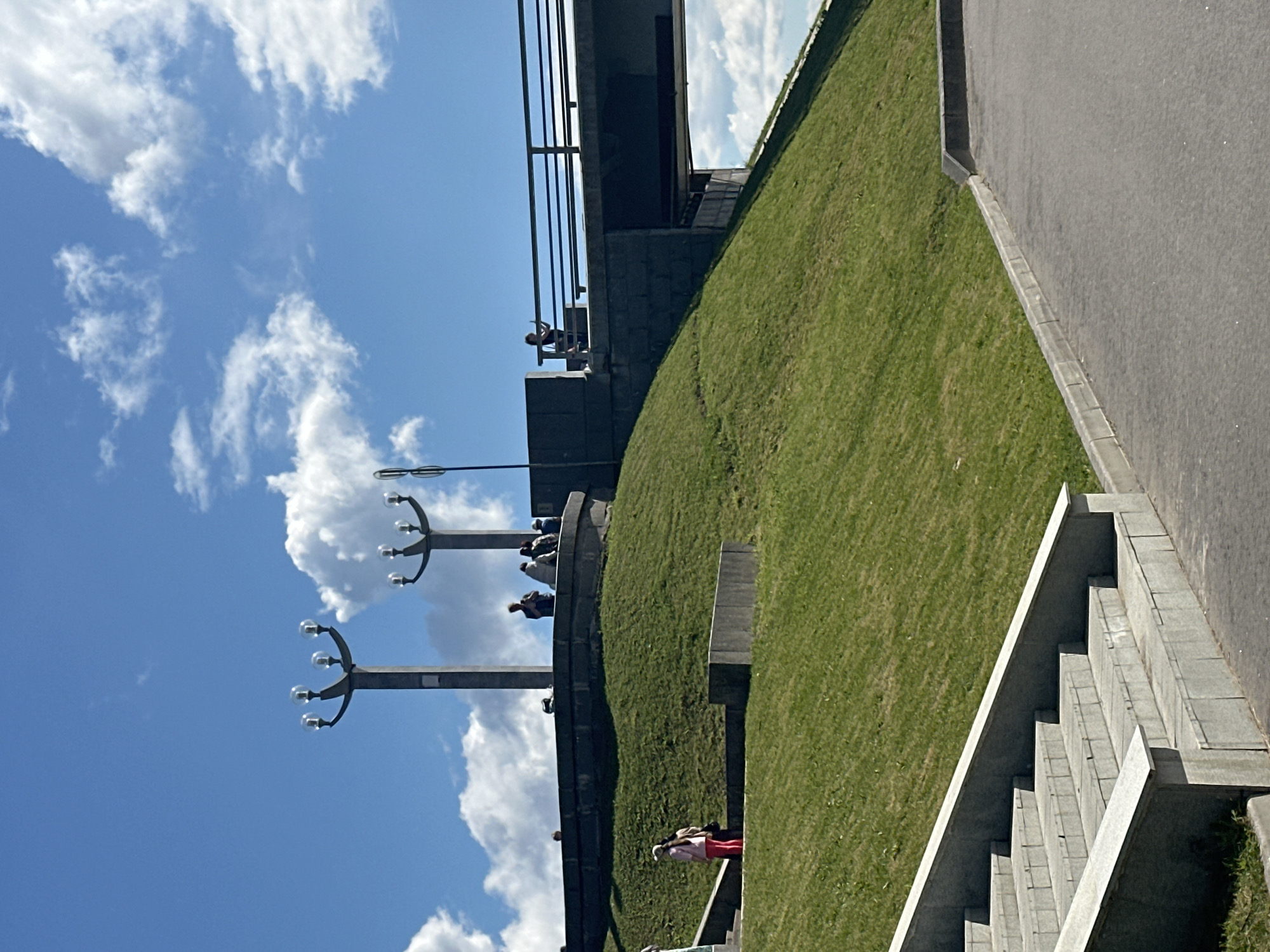 The image shows an outdoor scene with a bright blue sky and scattered white clouds. In the foreground, there is a set of wide concrete stairs. To the right of the stairs, a paved area leads away. Behind the stairs, a grassy embankment rises, with two people standing near its base on the left. The embankment is topped by a dark, curved concrete structure with several people seated and standing on it. Two ornate street lamps with multiple bulbs are positioned on this upper level. Further to the right, a concrete overpass or bridge crosses the scene. A single person is visible standing on the railing of this bridge. The scene appears to be a public park or promenade in the city of Veliky Novgorod, Russia.