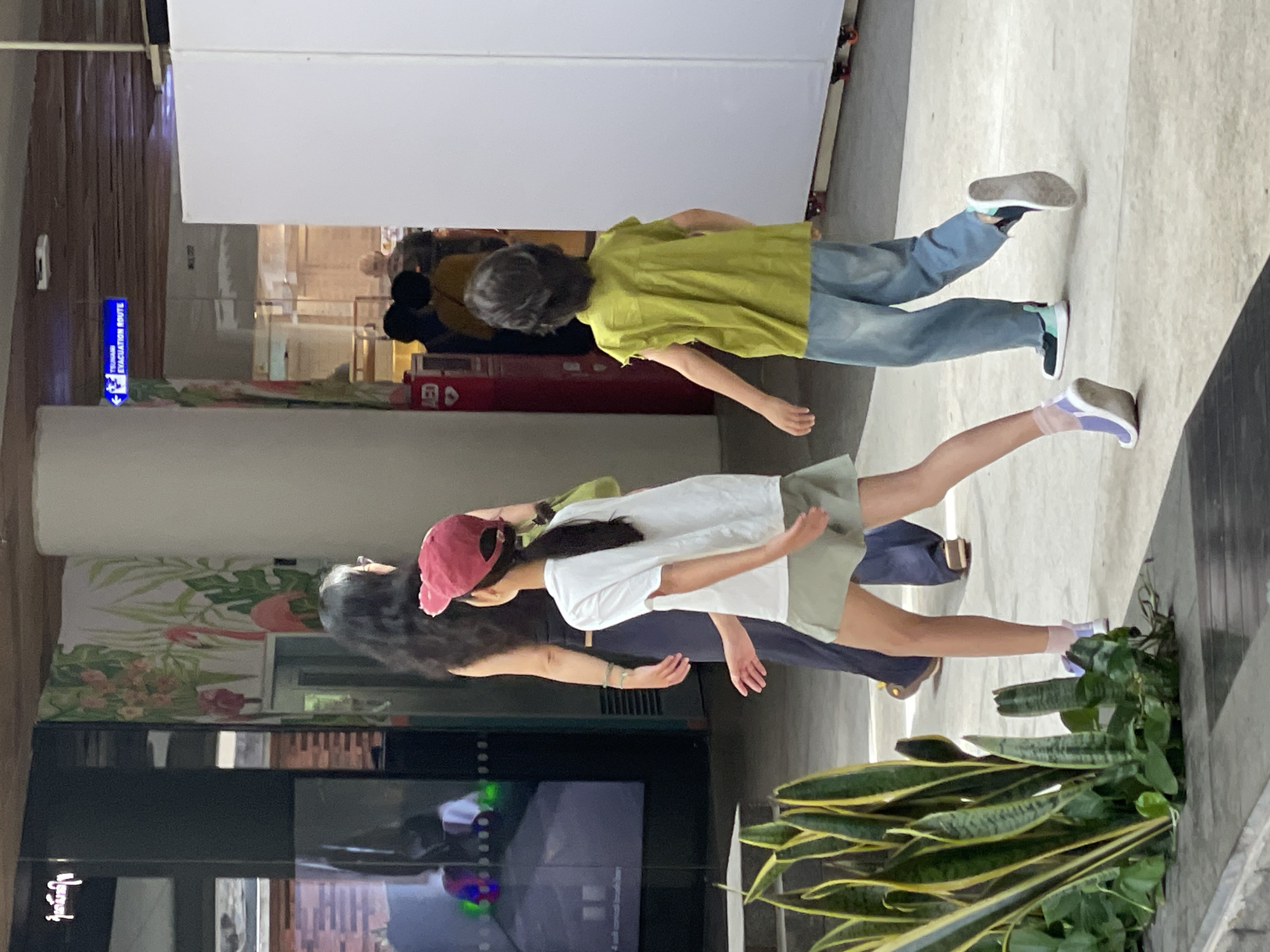 In Kuta, Indonesia, on a paved indoor walkway with natural light filtering in, three individuals are captured in motion. A woman with long dark hair, wearing a dark top and dark pants, walks away from the camera, holding a light green bag over her left shoulder. Ahead of her, a young girl in a white t-shirt and a light green skirt is also walking away, her body turned to her left. She wears a red baseball cap and white sneakers with light purple accents. To the right of the girl, another young person with dark, medium-length hair is walking away, wearing a lime green sleeveless shirt and light blue pants. They are also wearing sneakers, one dark with a light blue sole, and the other dark with a light blue accent. In the background, a large white display panel is visible, and to the left, there is a black display case with a screen showing abstract visuals and a large decorative panel with images of flamingos and tropical leaves. A blue sign indicating an "EMERGENCY EVACUATION ROUTE" is mounted on the ceiling. Lush green plants with long, striped leaves are situated in a planter box in the foreground, to the left of the frame.