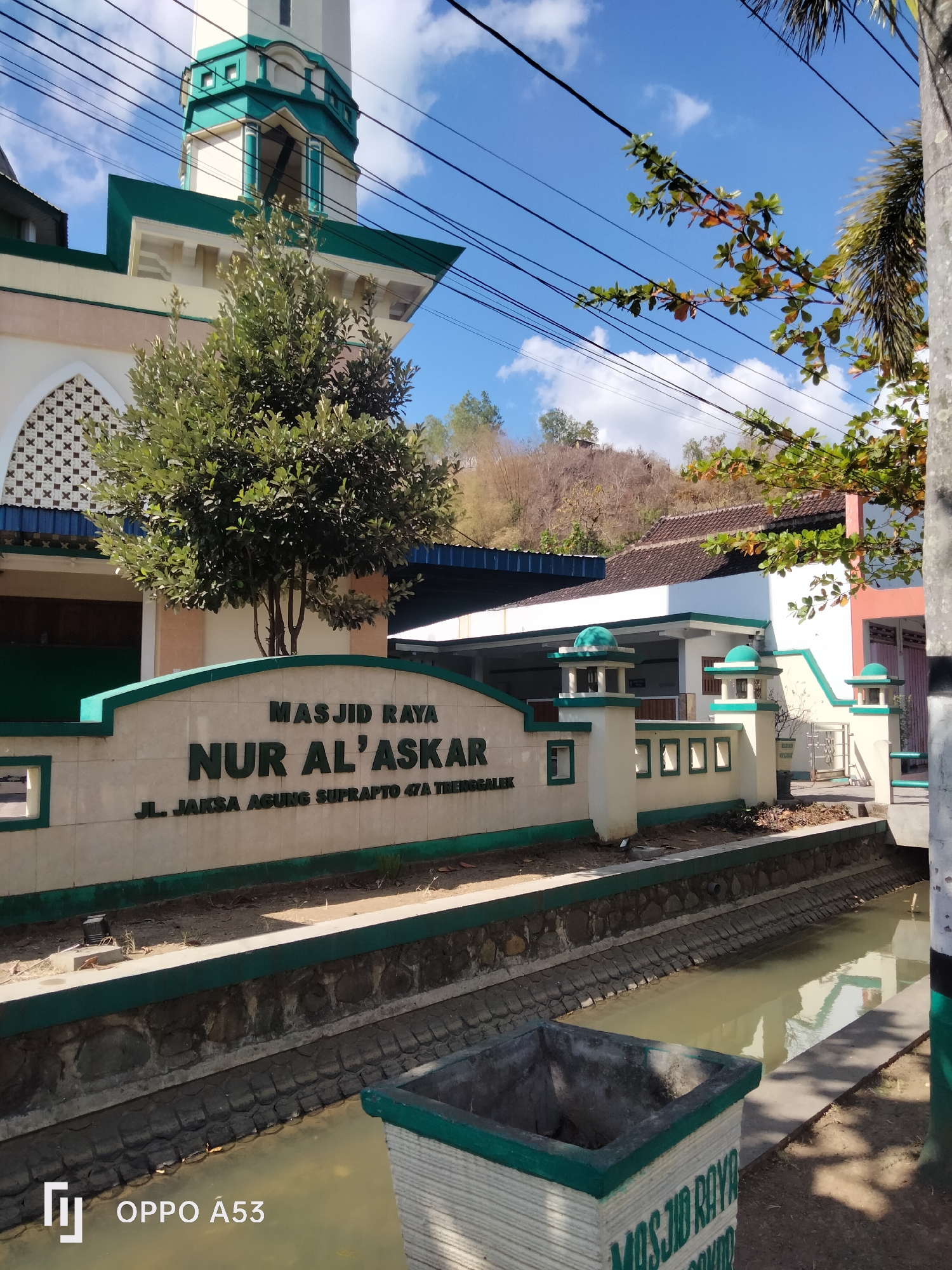 The image shows a facade of a mosque with a minaret against a bright blue sky with scattered clouds. In the foreground, there is a stone wall with the inscription "MASJID RAYA NUR AL'ASKAR" and below it "JL. JAKSA AGUNG SUPRAPTO 47A TRENGGALEK". A small tree is planted in front of the wall. To the right, a canal runs alongside the mosque, with a concrete planter box in the lower right corner. The overall scene depicts a sunny day at a mosque in Trenggalek, Indonesia.