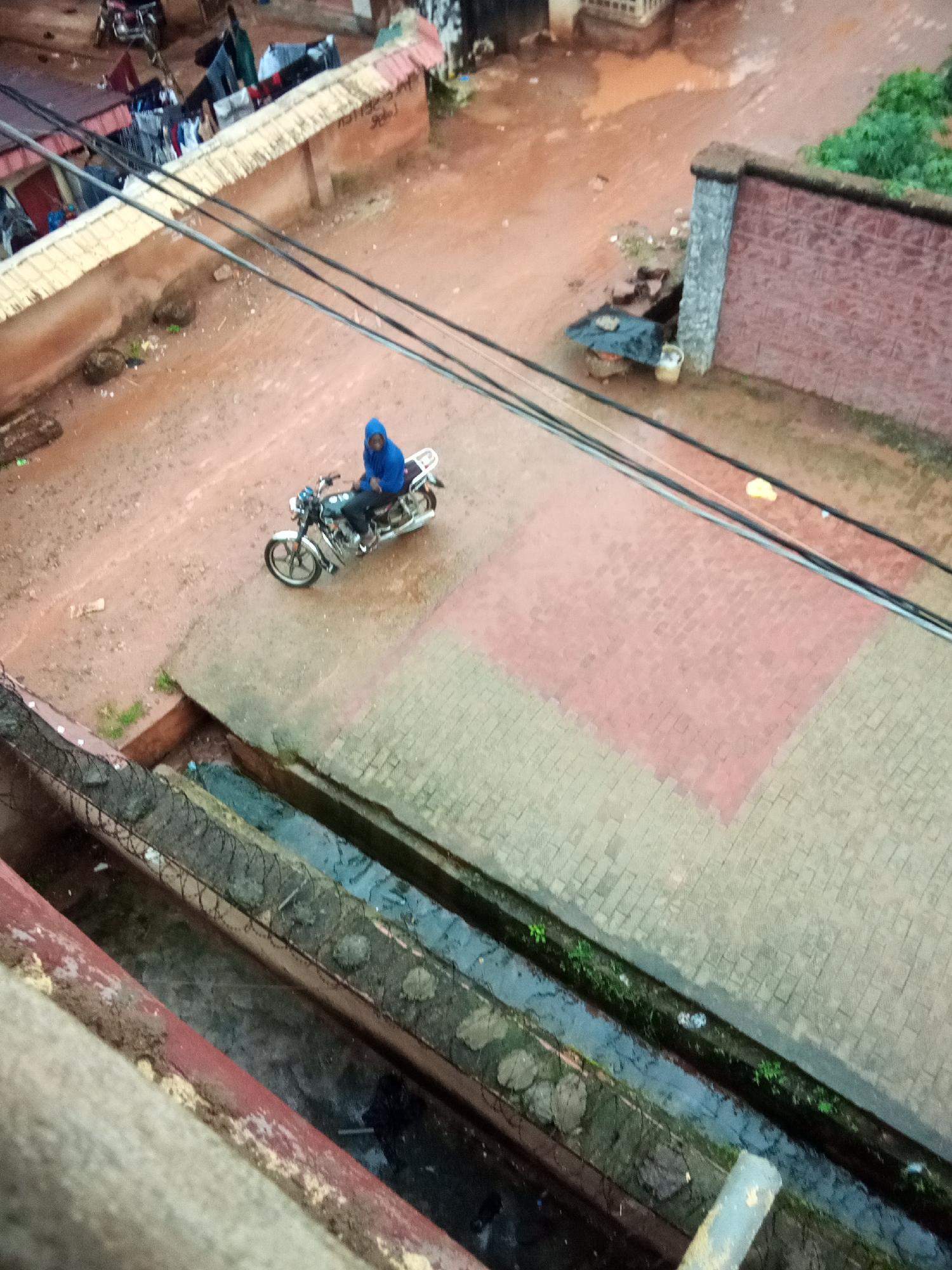 The image depicts an outdoor scene in Amaokpala, Nigeria. A person wearing a blue hooded garment is seated on a motorcycle, which is parked on a paved area. The ground is predominantly red earth and is wet in places, suggesting recent rain. Several power lines are visible crisscrossing the scene. In the background, there are buildings with corrugated iron roofs, and laundry is hung out to dry on lines. A stone wall with a brick pattern borders a section of the area. A narrow, concrete drainage channel filled with murky water runs along the foreground, bordered by a fence with barbed wire. The overall environment appears to be a semi-urban or rural residential area.