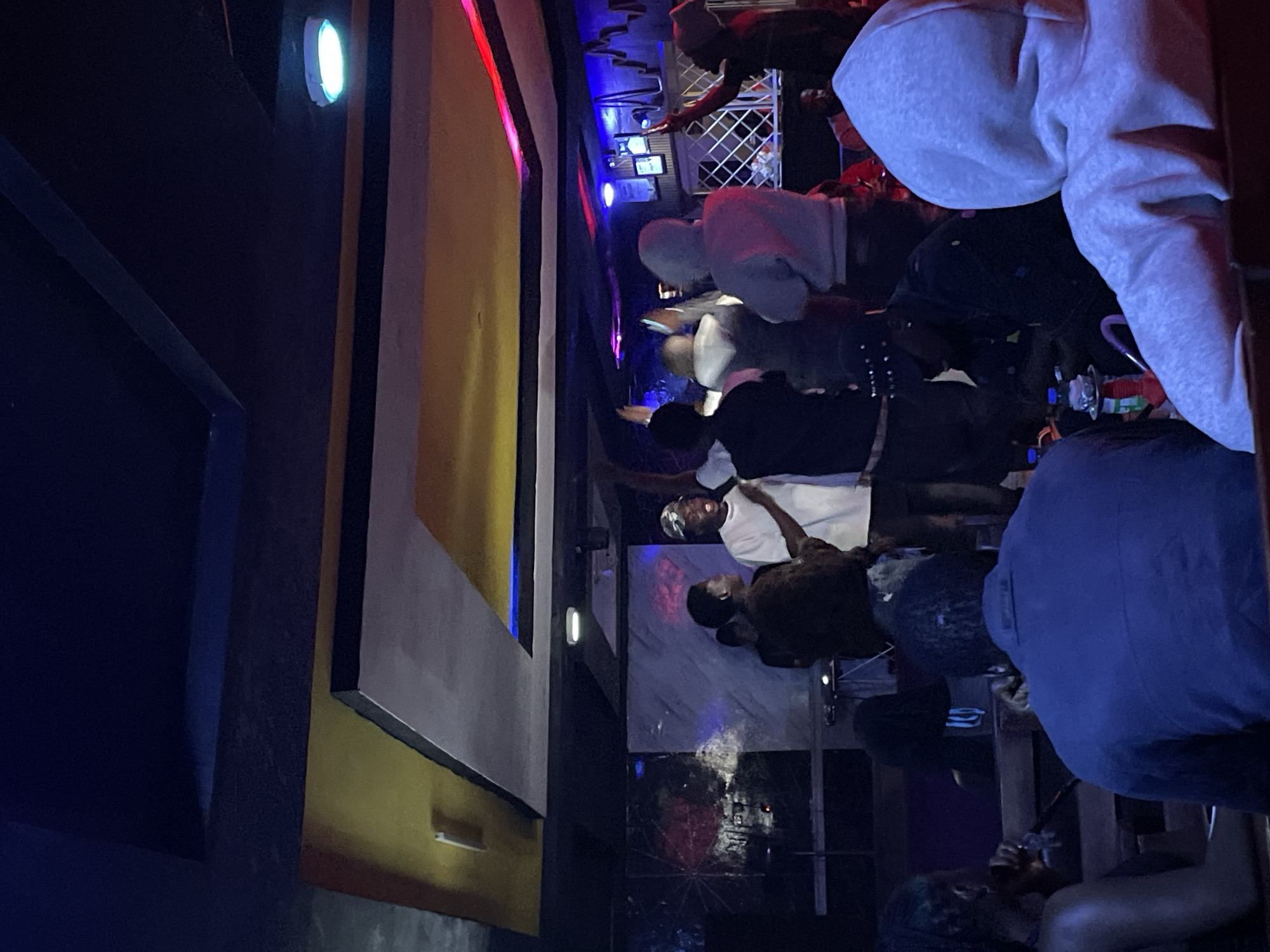 The image depicts an indoor gathering at a venue in Durumi, Nigeria. Several individuals are present, with a prominent group gathered around a stage area where a person in a white t-shirt appears to be performing or speaking into a microphone. The environment is dimly lit, with colorful lighting, including blue and red accents, contributing to a vibrant atmosphere. In the foreground, several people are seated, facing away from the camera, suggesting they are observing the activity on stage. One individual in the far foreground wears a light grey hooded sweatshirt. To the left, a person with green-tinted hair is visible. The background includes decorative elements on the walls and a ceiling with tiered sections, illuminated by overhead lights. The overall scene suggests a social event or performance taking place within a nightclub or similar entertainment venue.