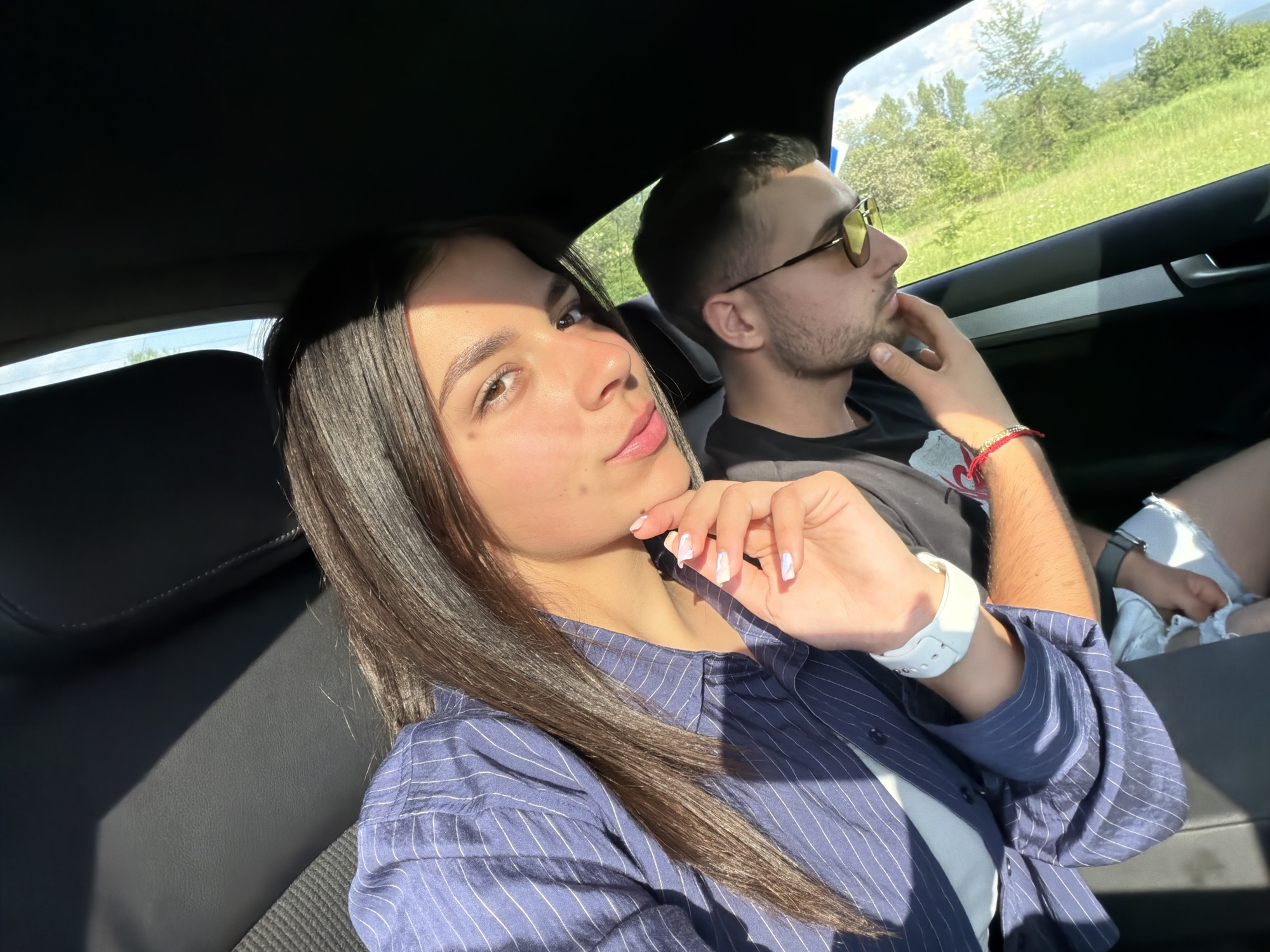 A couple is in a car. The woman in the foreground is wearing a blue striped shirt and a white watch, holding her hand near her face. The man in the background wears sunglasses and a black T-shirt, with a hand near his chin. They appear to be driving through a green, rural area. Given the location provided, the image may have been taken near Хаджийско, Bulgaria. The interior of the car is dark, with leather seats.