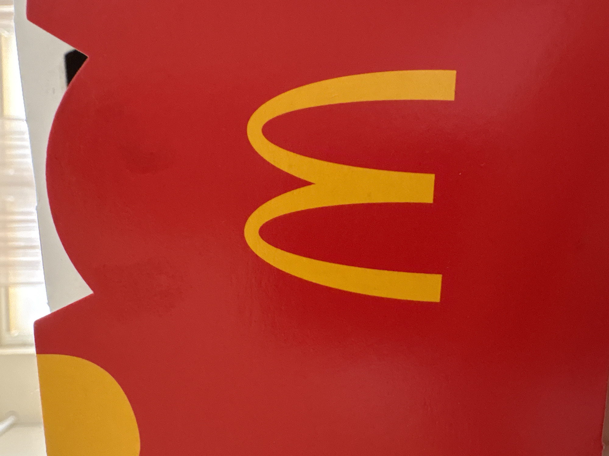 A close-up shows a red McDonald's Happy Meal box with a partially visible golden arches logo on the side. The box is positioned in front of a window with blinds. The location cannot be determined from the image.