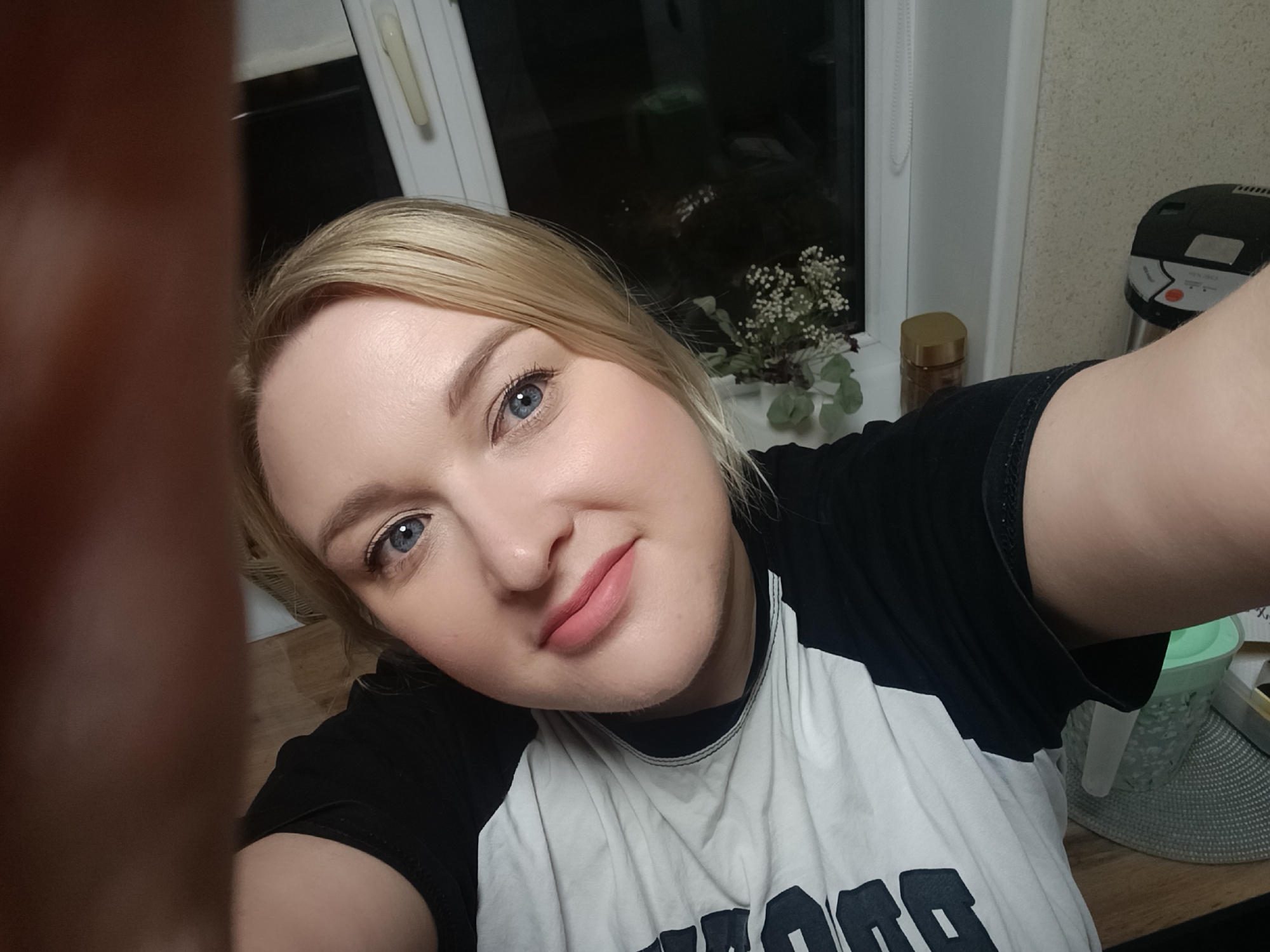 A blonde woman with blue eyes and light pink lipstick is depicted in a close-up selfie, smiling faintly. She wears a black and white raglan-style t-shirt with a partial dark graphic on the white torso section. Her right arm extends into the left foreground, indicating she is holding the camera. The background reveals an indoor setting, likely a kitchen or dining area. A white-framed window, showing darkness outside, is positioned behind her, suggesting nighttime. On the windowsill below the window, a small white potted plant with delicate white flowers and green foliage is visible, alongside a golden-lidded jar. To the right, a black and silver kitchen appliance (possibly a rice cooker) rests on a wooden-patterned countertop, with a grey textured placemat underneath it. A light-colored, subtly textured wall is visible to the right of the window. This scene is located in Chaikovsky, Russia.