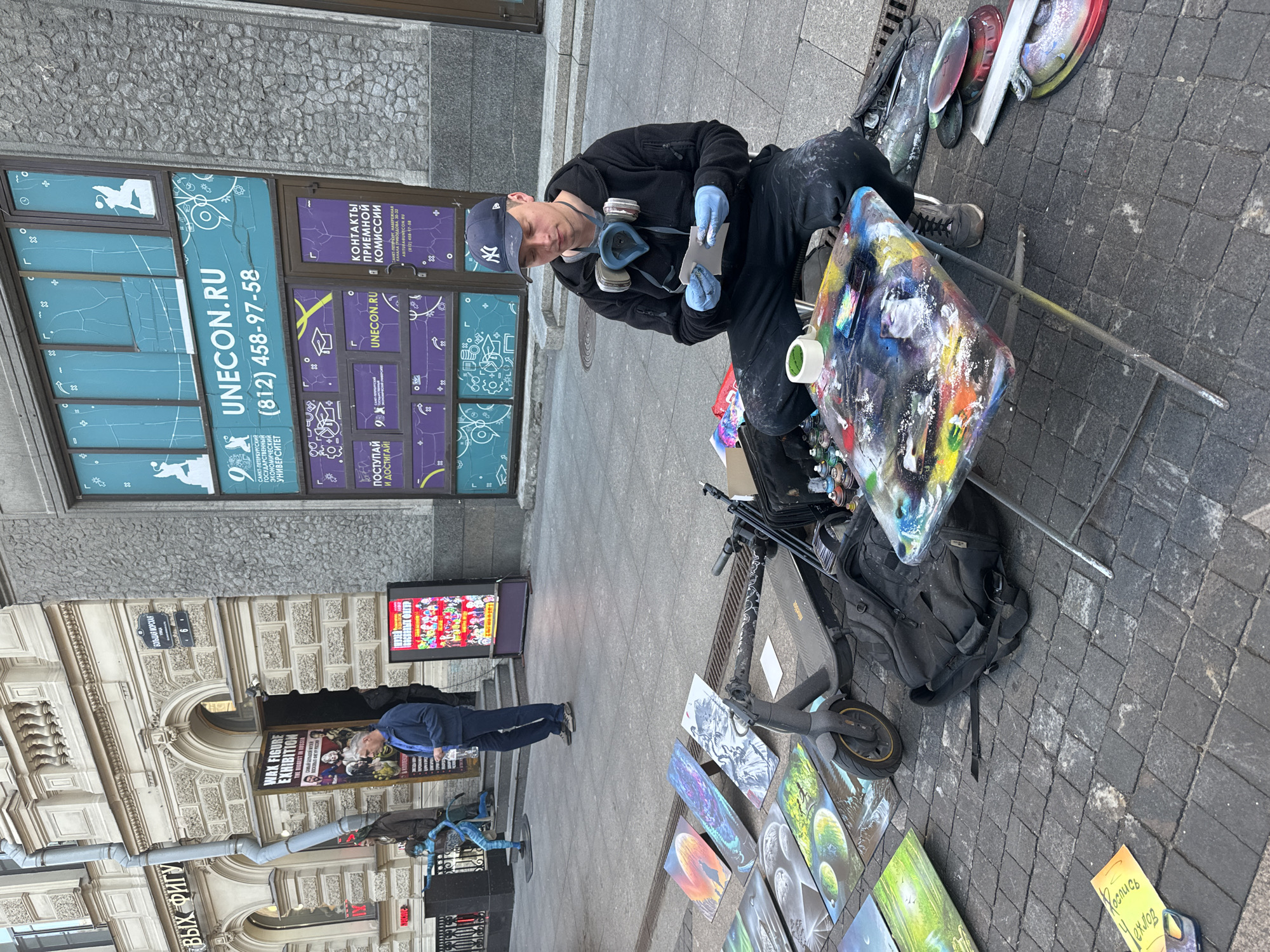 An artist, wearing a black hoodie, black pants, a blue New York Yankees baseball cap, a respirator mask, and blue gloves, sits on the sidewalk in Saint Petersburg, Russia, preparing to spray paint. Spread out before him are several colorful canvases, a black backpack, a dark gray electric scooter, and a portable table covered in spray paint cans and abstract designs. In the background, a building with large windows displays advertisements for "UNECON.RU" and the "Wax Figure Exhibition." A blue statue resembling the character "Neytiri" from the movie Avatar stands near the entrance to the exhibition. The scene takes place on a paved sidewalk with cobblestone detailing and a metal grate.