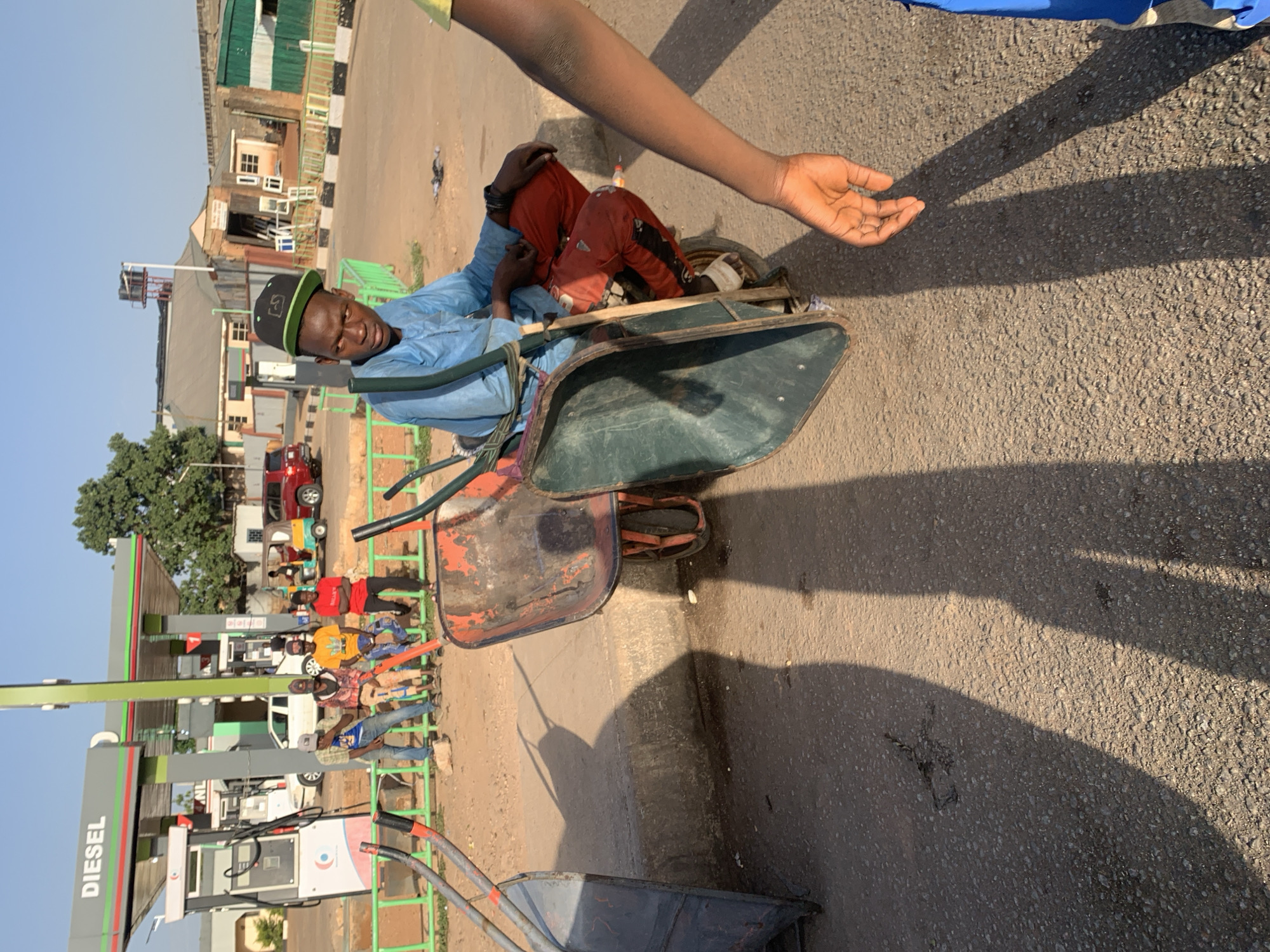 A man in a blue shirt and black cap sits in a green wheelbarrow on a paved road, with his legs inside the wheelbarrow, near a gas station in Gura Top, Nigeria. Another arm extends into the frame from the top right. Other wheelbarrows are nearby on the road. Several people sit near the gas station, and buildings are in the background.