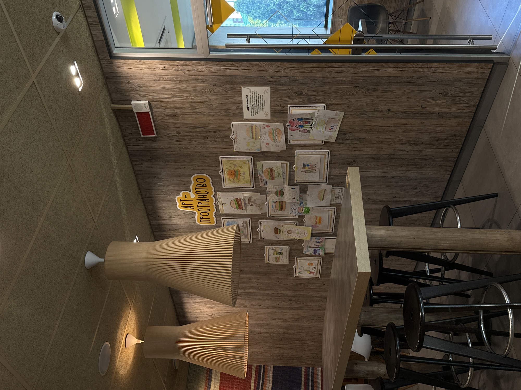 The image depicts the interior of a McDonald's restaurant in Minsk, Belarus, specifically the seating area with a long, light-wood table and black stools. The walls are paneled with wood grain and decorated with children's drawings, many featuring burgers, and a yellow sign with Russian text that reads "АРТ-ПРОСТРАНСТВО" (Art Space). Two large, cone-shaped wooden lamps hang from the beige acoustic tile ceiling. To the right, a glass door leads to another area of the restaurant, with a glimpse of an exterior window showing trees. A security camera is visible on the ceiling near the upper right corner. The overall atmosphere is casual and family-friendly, typical of a fast-food establishment.