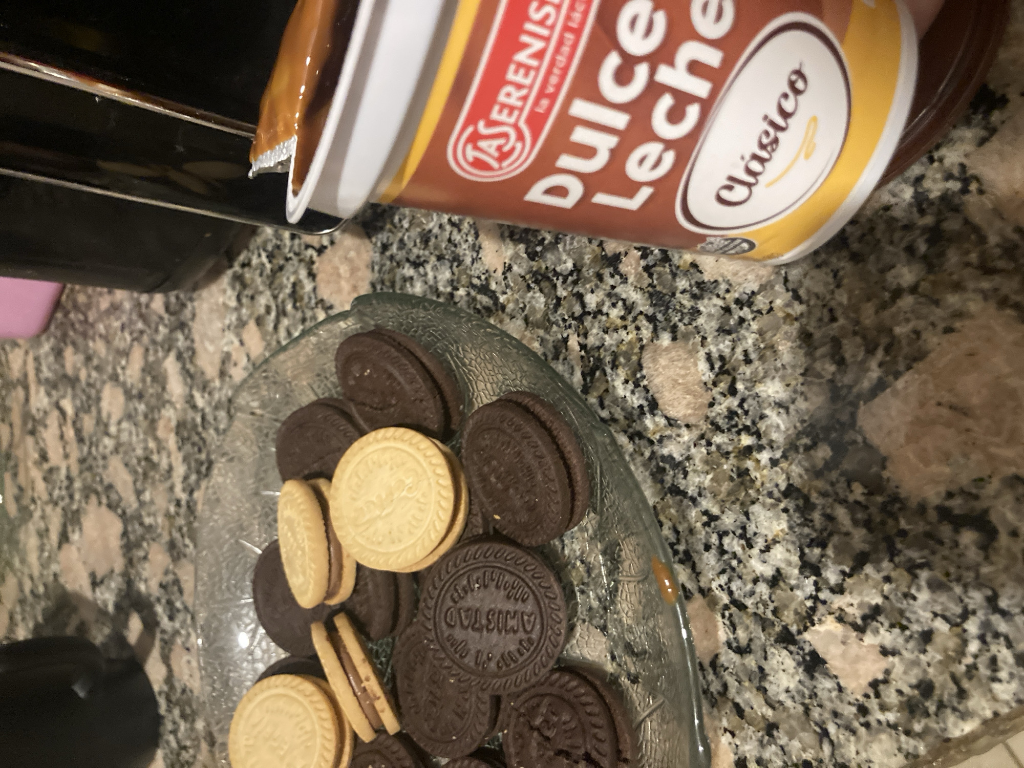 The image captures an indoor scene, likely a kitchen, with a speckled granite countertop serving as the primary surface. A large container of "LA SERENISIMA DULCE de LECHE Clásico" is prominently positioned in the upper right, open and angled as if being used or poured. A small amount of the caramel-colored dulce de leche is visible near the container's opening, and a slight smudge of it is on the countertop near a glass plate.
The glass plate or bowl, situated in the center-left, holds an assortment of sandwich cookies. These cookies come in two main varieties: some are light-colored with cream filling, while others are dark-colored with a similar filling. Several of the dark cookies have the word "AMISTAD" embossed on their surface.
In the upper left background, part of a dark-colored appliance or object is visible. Further in the background, a pink container can be discerned on the far left. A black object or container also appears in the lower left corner. The overall scene suggests the preparation or enjoyment of a snack, possibly involving dipping the cookies in the dulce de leche. No people are visible.