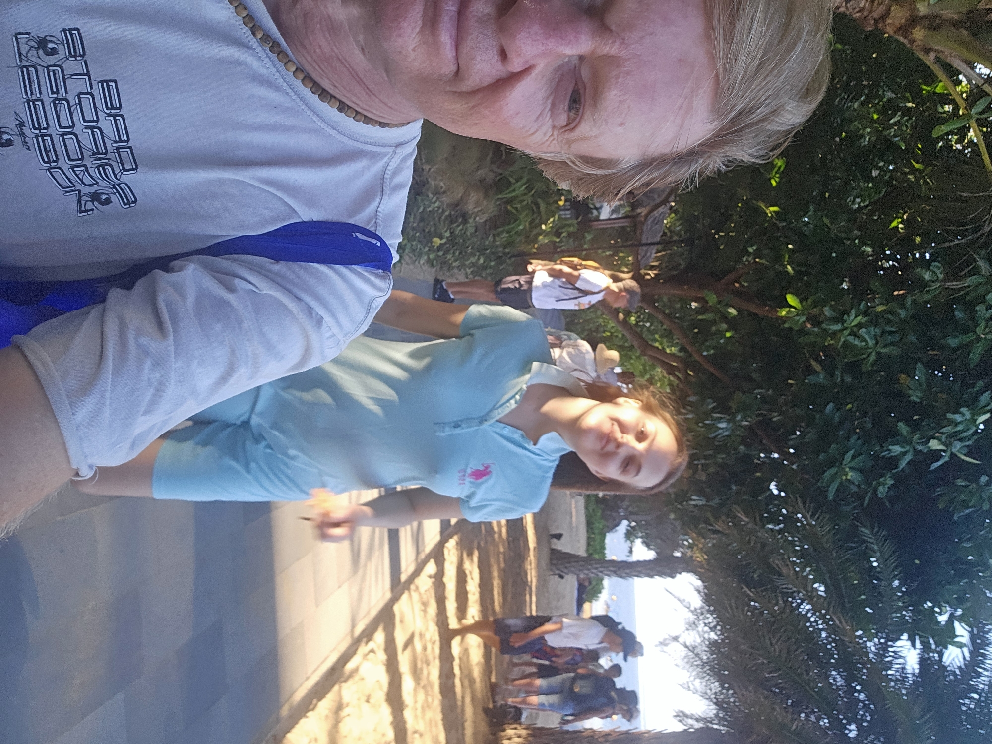The image captures a selfie taken by a man with fair skin and blond hair, wearing a light-colored t-shirt adorned with a graphic design and a wooden beaded necklace. To his right, a woman with brown hair is walking away, wearing a light blue polo shirt. In the background, a sandy path is visible, lined with palm trees and lush green foliage. Several other individuals, including men and women, are scattered along the path, some appearing to be tourists. The scene is set outdoors, likely in a tropical environment, possibly along a beach or promenade. The overall atmosphere suggests a casual outing in a warm climate. The location is described as Denpasar, Indonesia, though no specific landmarks are identifiable in the image to confirm this.