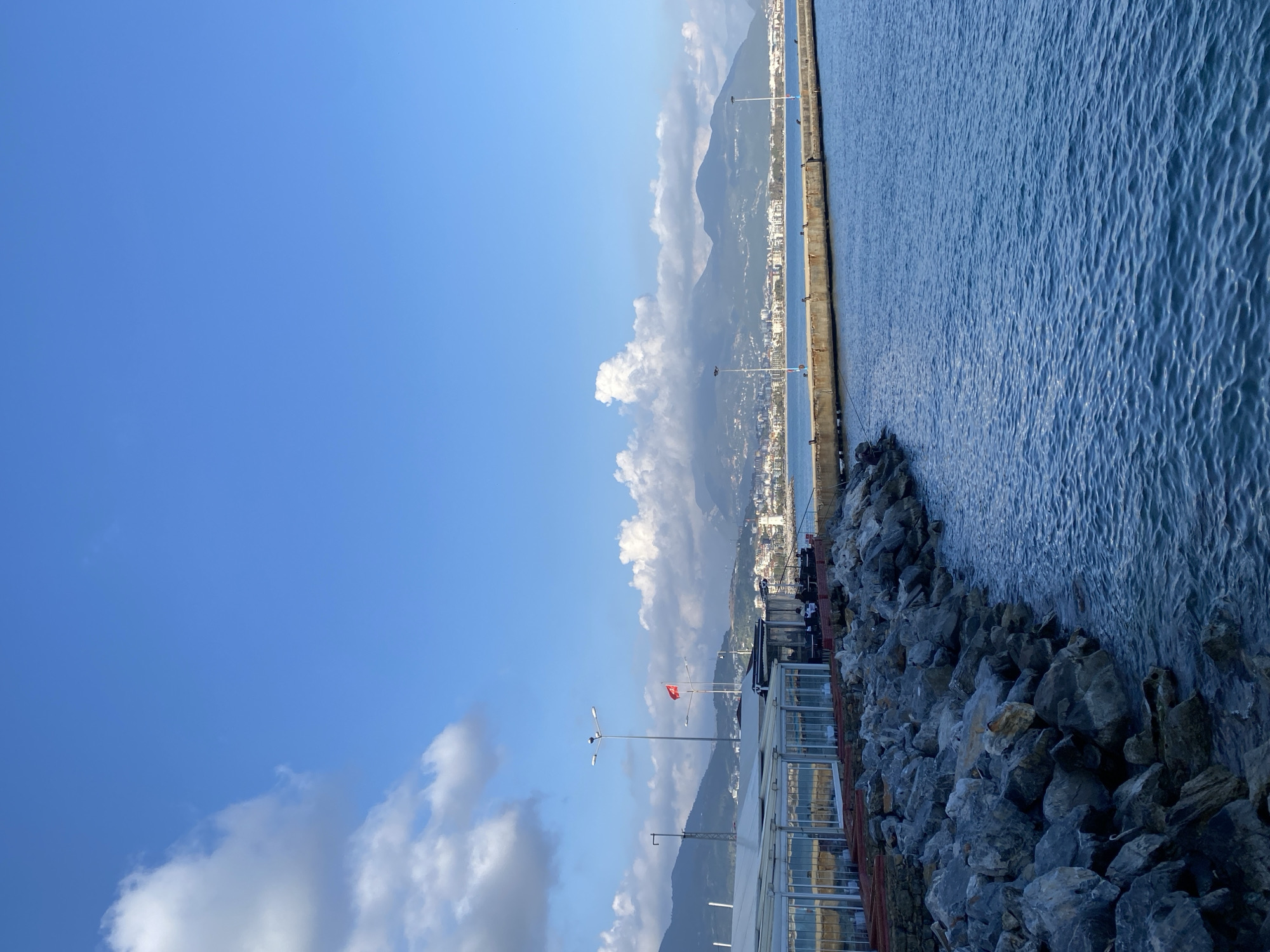 This image captures a vibrant coastal scene under a clear blue sky with scattered white cumulus clouds. A vast expanse of blue water, likely the Mediterranean Sea, dominates the right side of the frame, exhibiting gentle ripples across its surface.
A long, straight concrete pier or breakwater extends into the distance, separating the open water from a more sheltered area closer to the shore. Along the left edge of the water and in the foreground, a rugged rocky shoreline or an extension of the breakwater is visible, composed of large grey boulders.
In the middle ground, a bustling coastal city or town stretches along the base of imposing, hazy mountains. Numerous buildings, some with reflective glass windows, make up the urban landscape. Several streetlights are positioned along the pier and near the coastal structures. A prominent red flag, clearly identifiable as the Turkish flag with its white star and crescent, flies from a pole in the mid-ground.
Indications of activity include a couple of fishing rods visible on the rocky shoreline in the foreground, suggesting people may be fishing, although individuals are not distinct.
The scene depicts a bright and sunny day, likely during the morning or late afternoon, given the clear conditions and soft light. The overall atmosphere is peaceful and scenic. The location is contextualized as Alanya, Turkey, which is consistent with the visible Turkish flag and coastal mountainous landscape. No discernible text is visible in the image.