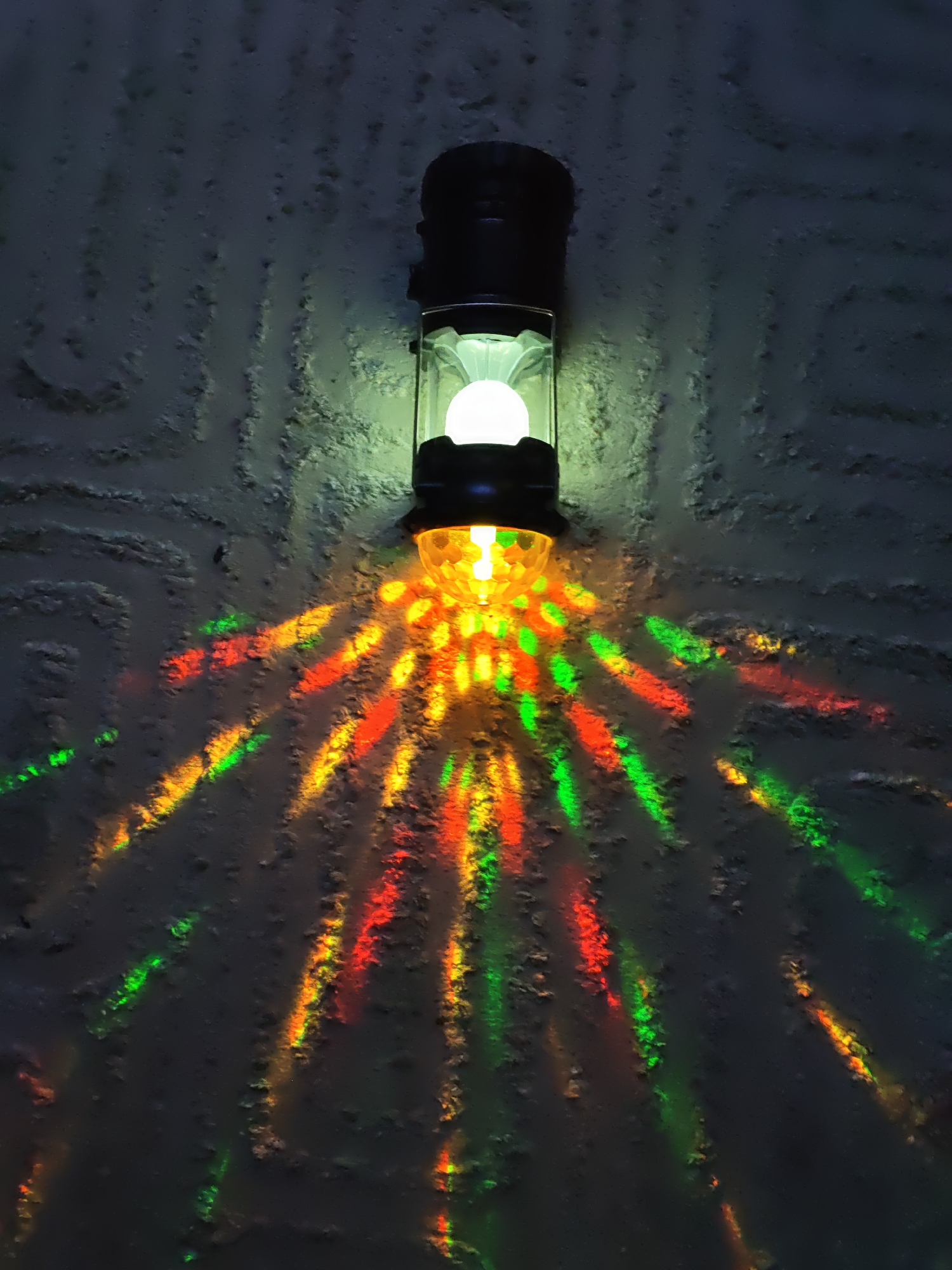 An illuminated lantern, mounted on a rough, textured wall, casts colorful light patterns. The lantern is black and clear with a white bulb inside, emitting green and yellow light, while below it, a faceted yellow globe projects red, orange, and green light in streaks across the wall. The wall itself is a dark, uneven surface, with vertical lines of texture and shadow. The image appears to be an interior shot, possibly in a dimly lit room or hallway, given the darkness of the background and the strong illumination from the lantern. There are no people or animals visible in the image. The scene is static and focuses on the interplay of light and shadow on the textured surface.