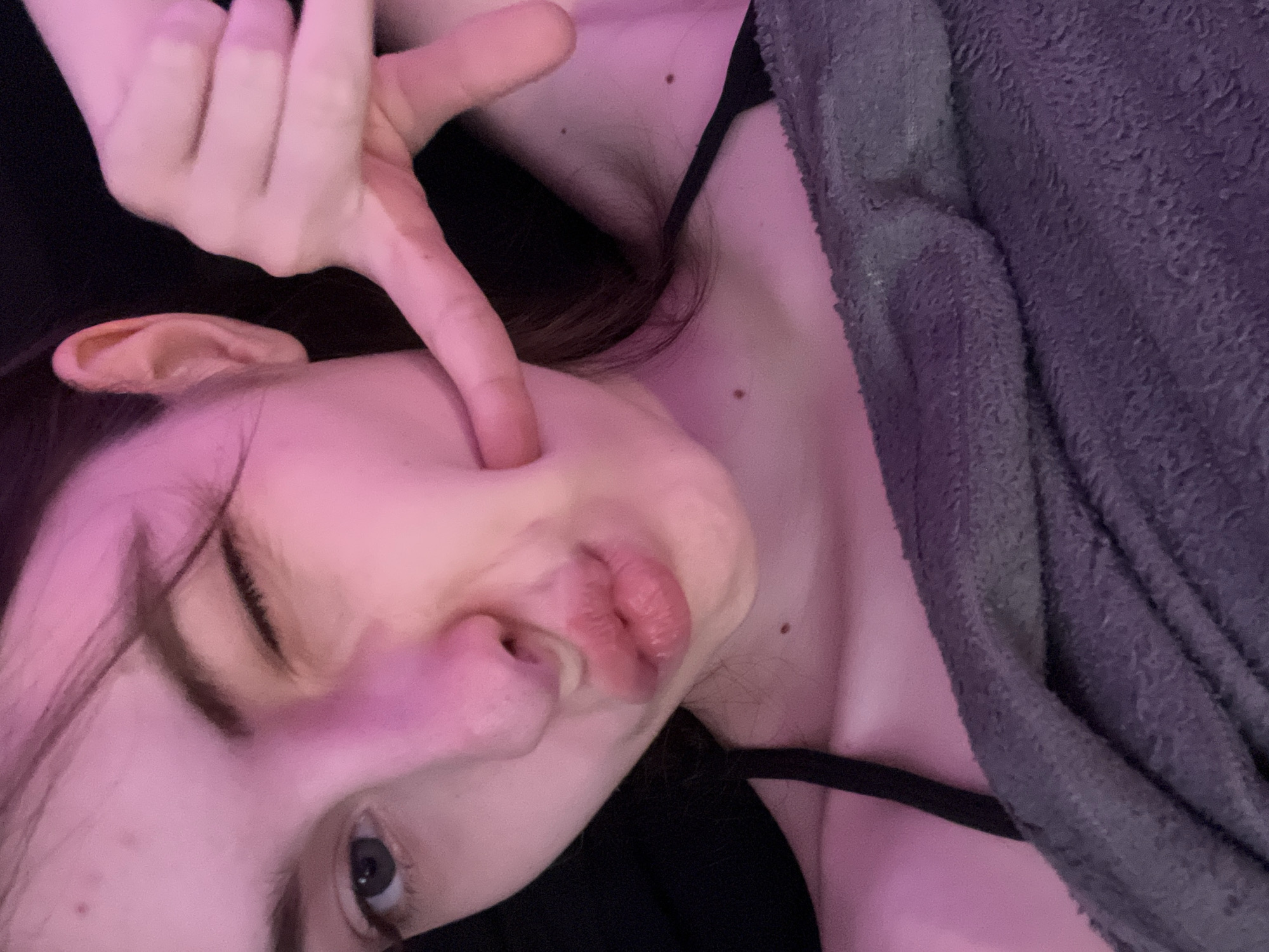 The image shows a close-up, angled view of a person lying down, possibly on a black surface. The person's face is prominent, with their cheek being pushed in by a finger, distorting their features. One eye is visible and open, while the other is closed. The person's lips are pursed. The person is wearing a thin, black strap garment. A gray, textured item, possibly a blanket or towel, is draped over the right side of the person. There are two moles visible on the person's neck and chest. The lighting appears to be artificial, with a pinkish hue. The location of the image is unknown and cannot be determined from the visual information.
