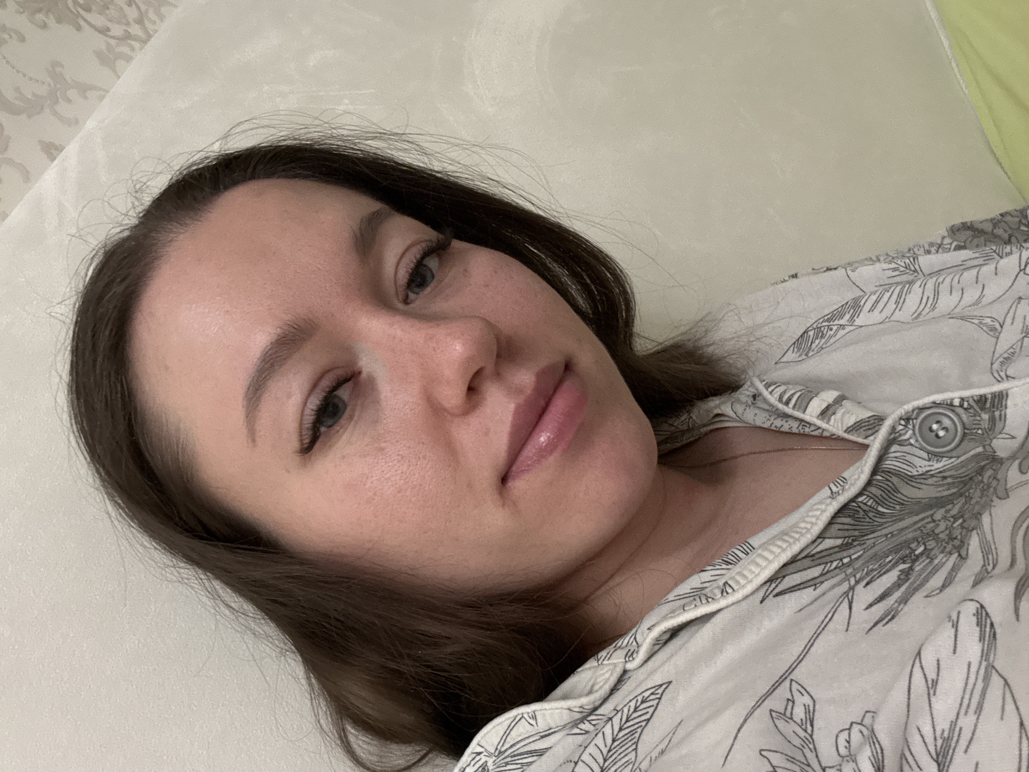 A woman lies on a light-colored surface, possibly a bed, in what appears to be an indoor setting. She has light skin, dark brown hair, and is wearing a patterned shirt. Her eyes are open, and she is looking at the camera. The location appears to be residential, but there are no distinct landmarks visible to confirm it is in the city of Ферма, Russia.