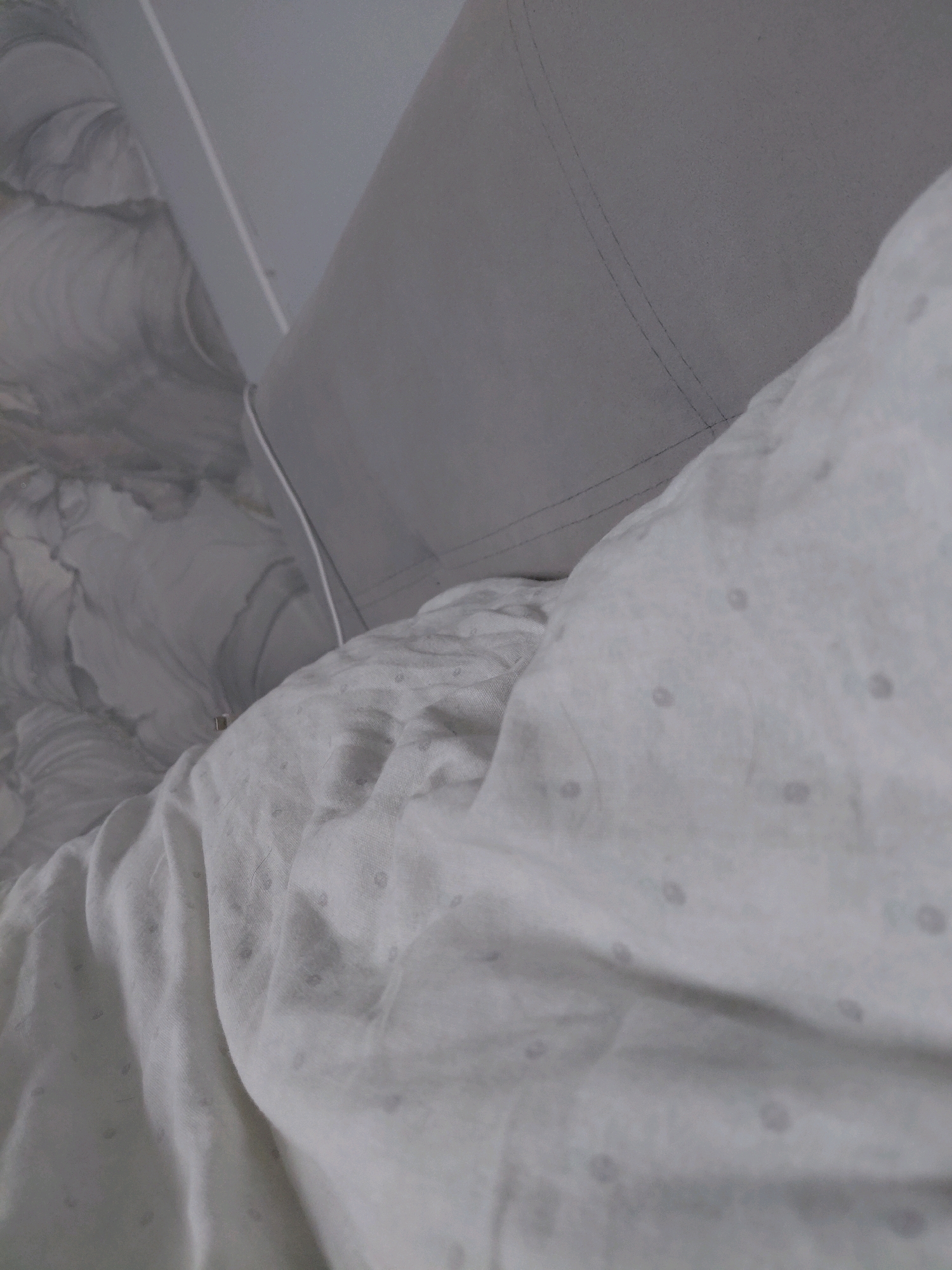 The image displays a close-up, oblique view of bedding. The foreground features a rumpled white sheet with a subtle, repeating pattern of small, dark dots. Behind this, a grey pillow or duvet is visible, characterized by straight stitching lines that suggest a structured form. Further in the background, a marbled-patterned fabric, possibly a blanket or duvet cover, is partially seen. The overall lighting is soft and diffused, creating a muted color palette dominated by whites and greys. There are no people or visible actions taking place, presenting a static and domestic scene. The location in Malinovka, Russia, is a contextual detail not visually ascertainable from the image itself.