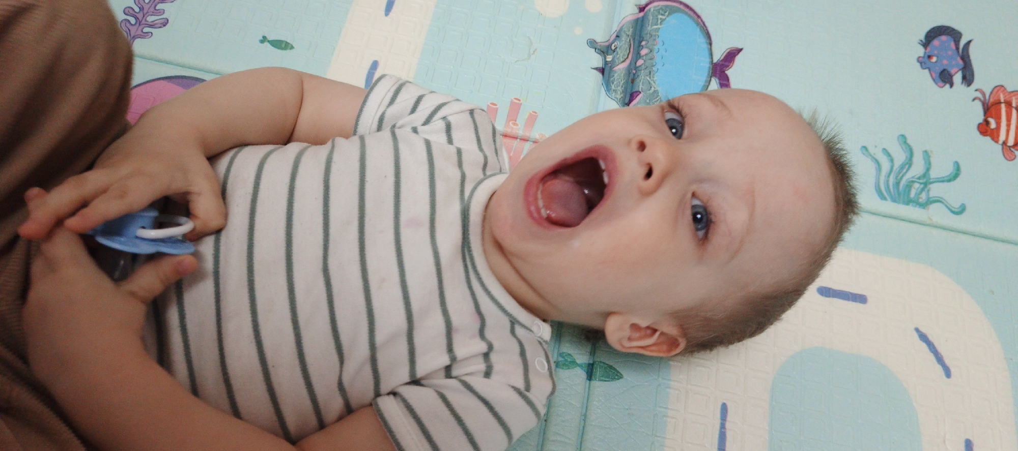 An overhead shot captures a baby lying on their back on a vibrant, light blue playmat adorned with colorful cartoon sea creatures like fish and coral. The baby, appearing to be an infant or young toddler, has fair skin, bright blue eyes, and light brown hair. They are dressed in a white onesie with thin, dark green horizontal stripes.
The baby's mouth is wide open, revealing their tongue and gums, suggesting they are either vocalizing loudly, expressing surprise or excitement, or perhaps mid-yawn. A small amount of drool is visible near the corner of their mouth. Their gaze is directed upwards towards the camera. In their left hand, the baby holds a light blue pacifier or teether with a white ring. The playmat's design includes various blue and white shapes, possibly depicting paths or water elements. The scene takes place indoors, likely during the daytime, in a child-friendly environment in Chaikovsky, Russia. There is no visible text in the image.