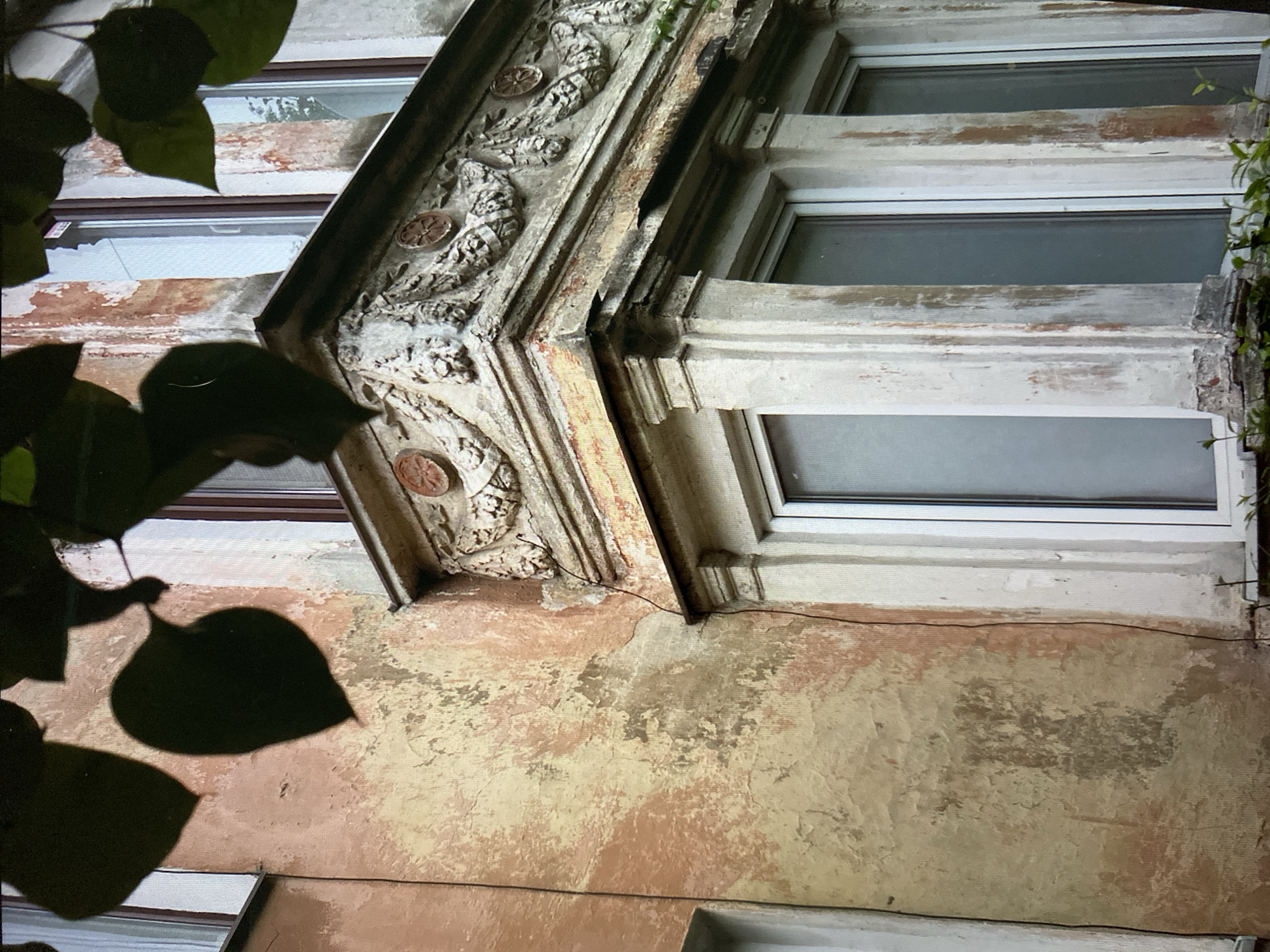 The image is a medium shot of a corner of an old building with peeling paint, located in the city of Khimki, Russia. The building features ornate decorative elements above a set of windows. The facade is weathered, with patches of light pink and yellow paint showing through areas of wear and tear. Dark green leaves from an overhead tree cast shadows on the building. The windows appear to be modern replacements in older frames. The overall impression is one of aging architecture in an urban setting.