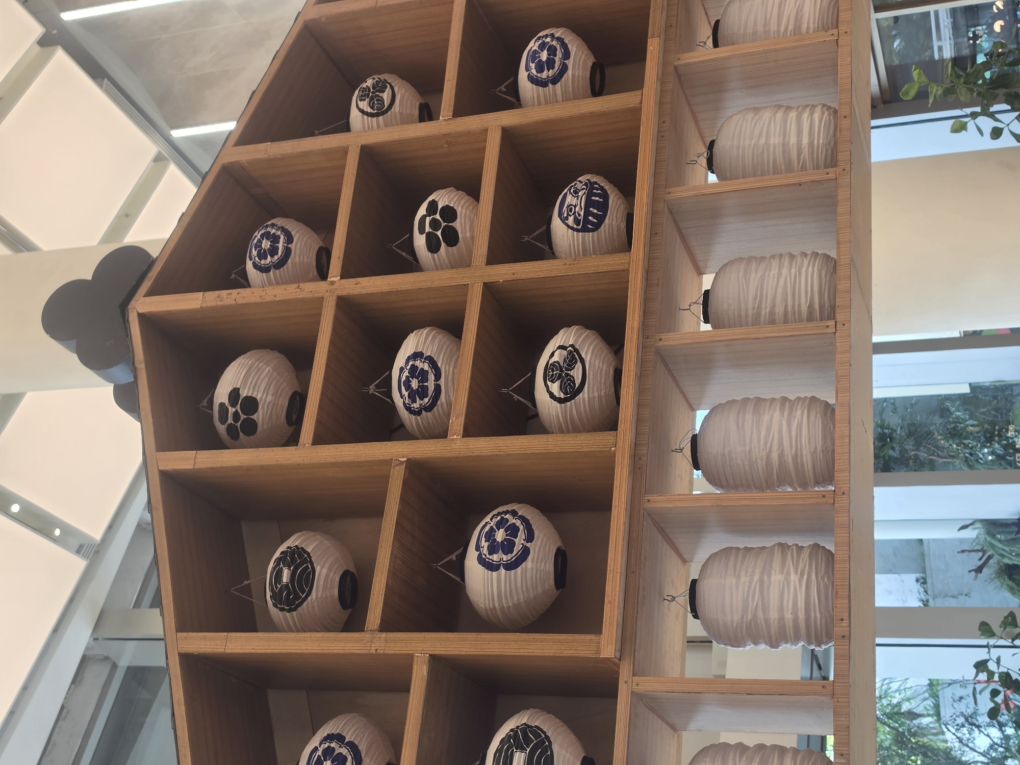This eye-level shot captures a decorative wooden shelving unit filled with white paper lanterns, creating an orderly and culturally themed display. The primary subjects are numerous spherical and cylindrical lanterns arranged within a light-colored wooden framework.
On the left and central sections, spherical or oval white paper lanterns are housed in individual square compartments. Many of these lanterns are adorned with distinct Japanese-style crests (mon), primarily in blue and black. Examples include a stylized floral pattern (possibly a chrysanthemum or plum blossom) and other geometric or organic motifs. These lanterns are held in place by small wire supports.
To the right, a separate section of the wooden unit contains elongated, cylindrical white paper lanterns. These lanterns are plain, unadorned, and have a ribbed texture. They are also supported by wires and are displayed horizontally within their rectangular compartments.
The wooden shelving unit itself is made of light, possibly untreated or lightly stained wood, featuring a grid-like structure with various sized compartments. In the upper left corner, a dark, cloud-like cutout element is partially visible, attached to the wooden structure.
Through large windows on the right side of the image, green foliage and outdoor elements are discernible, suggesting a bright daytime setting. Overhead, modern strip lighting fixtures are visible on a white ceiling. The overall scene suggests an indoor public space, likely a Japanese-themed establishment such as a restaurant or cultural center in Bangkok, Thailand. There is no visible activity or text in the image.